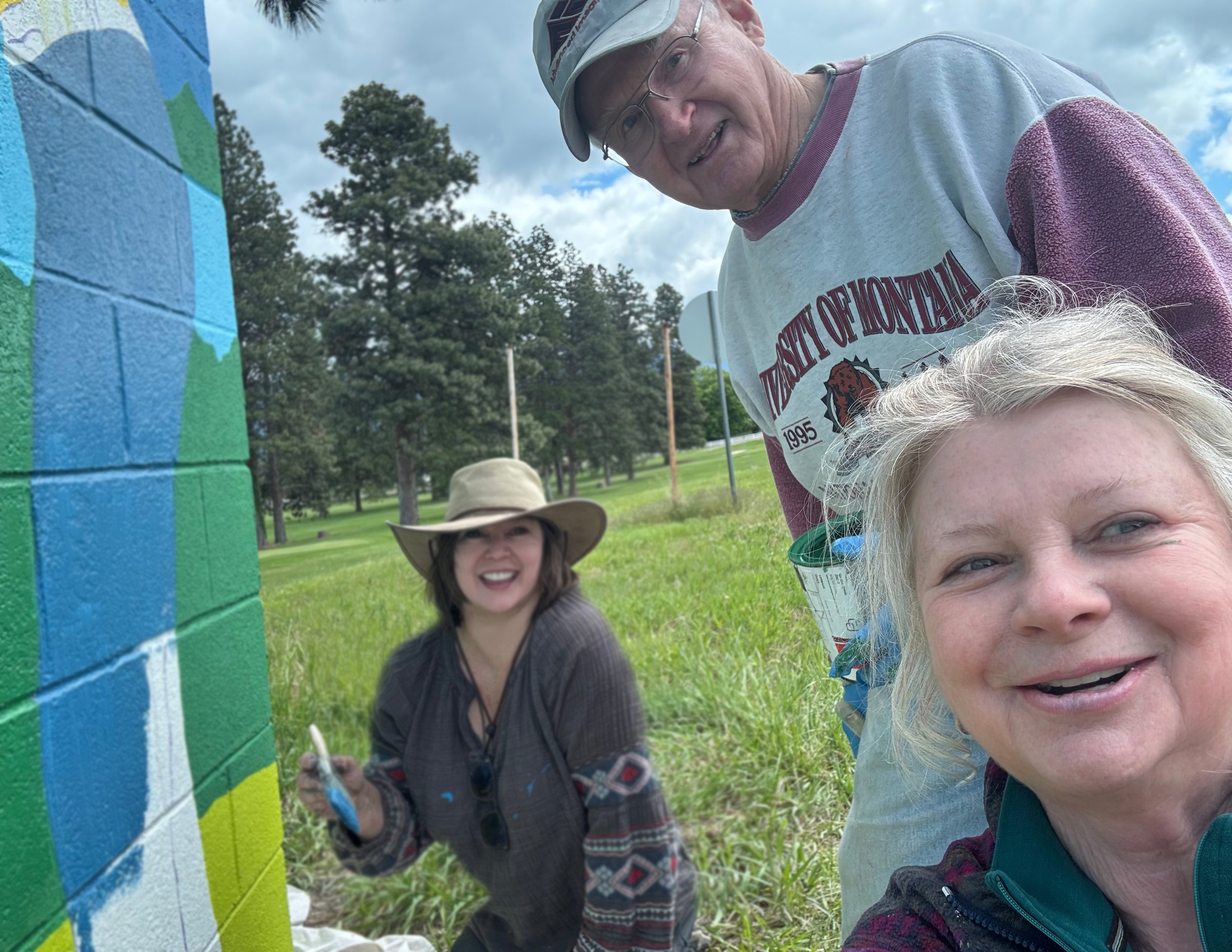 Three people outdoors, painting a large brick wall with bright colors. One person is holding a paintbrush and wearing a wide-brimmed hat, another is smiling with short light-colored hair, and the third is wearing glasses and a baseball cap. The scene includes green grass, trees, and a cloudy sky.