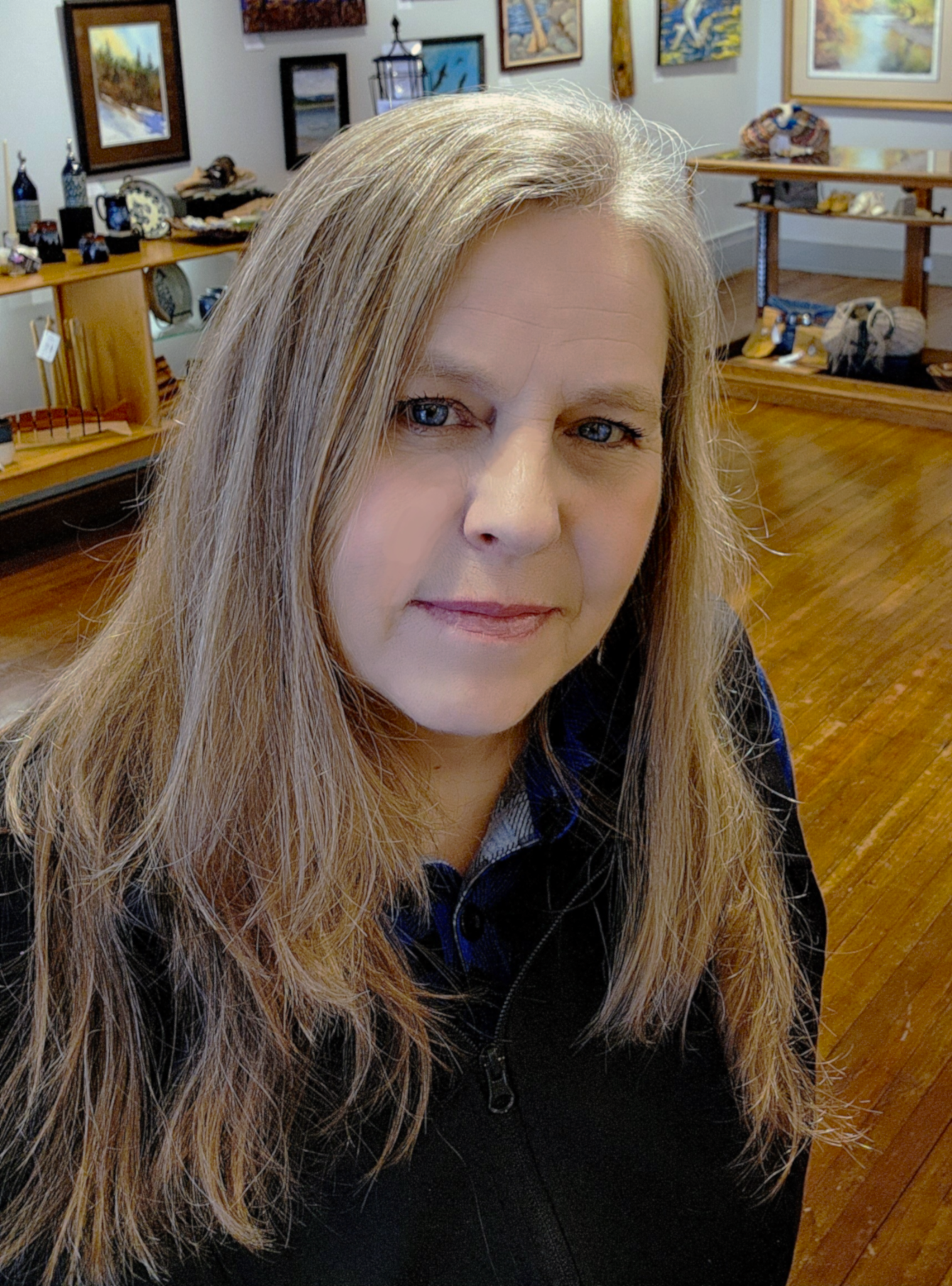 Close-up of a woman with long blonde hair and blue eyes, wearing a black jacket, in an art gallery or gift shop with paintings and decorative items on shelves in the background.