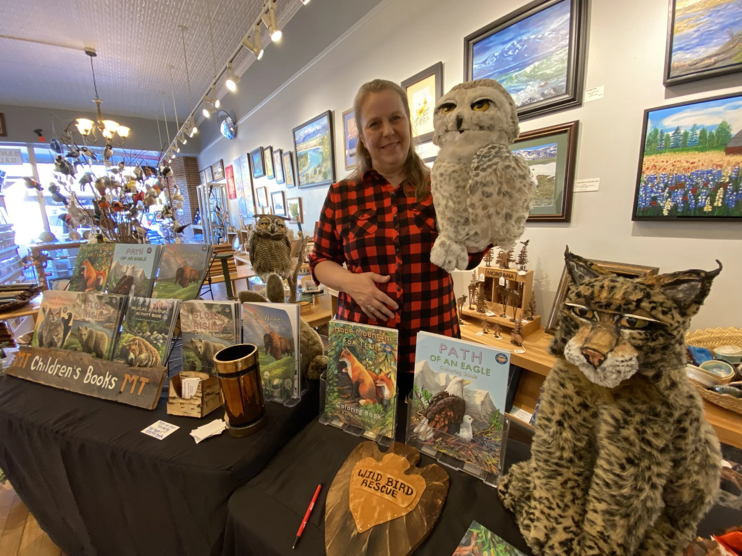 Karla Martinson standing at table with her animal friends, children's books, and artwork displayed in art gallery.