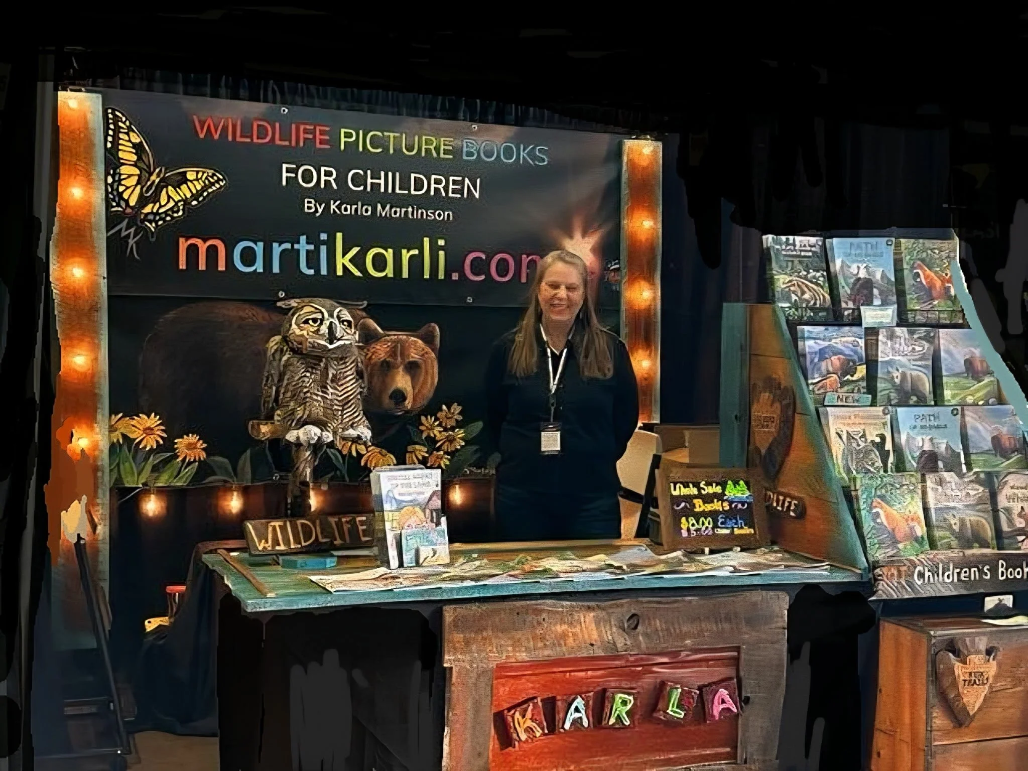 A woman standing behind a booth at a book fair, promoting wildlife picture books for children, with displays of books and animal-themed decor including a butterfly, an owl, a bear, and flowers.