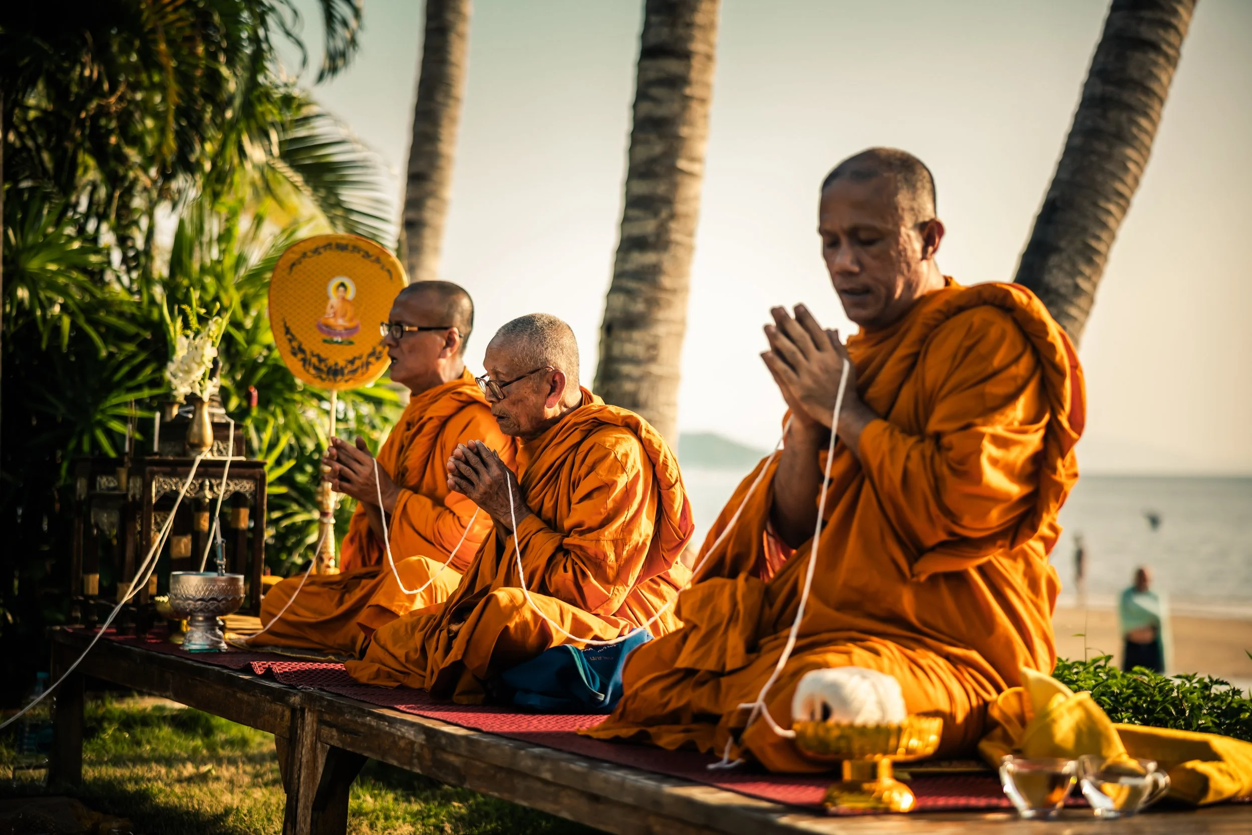 Four Buddhist monks wearing orange robes are praying on a bench outdoors near palm trees, with the ocean in the background. They are sitting with their hands in prayer, accompanied by religious items and an orange fan with an image of Buddha.