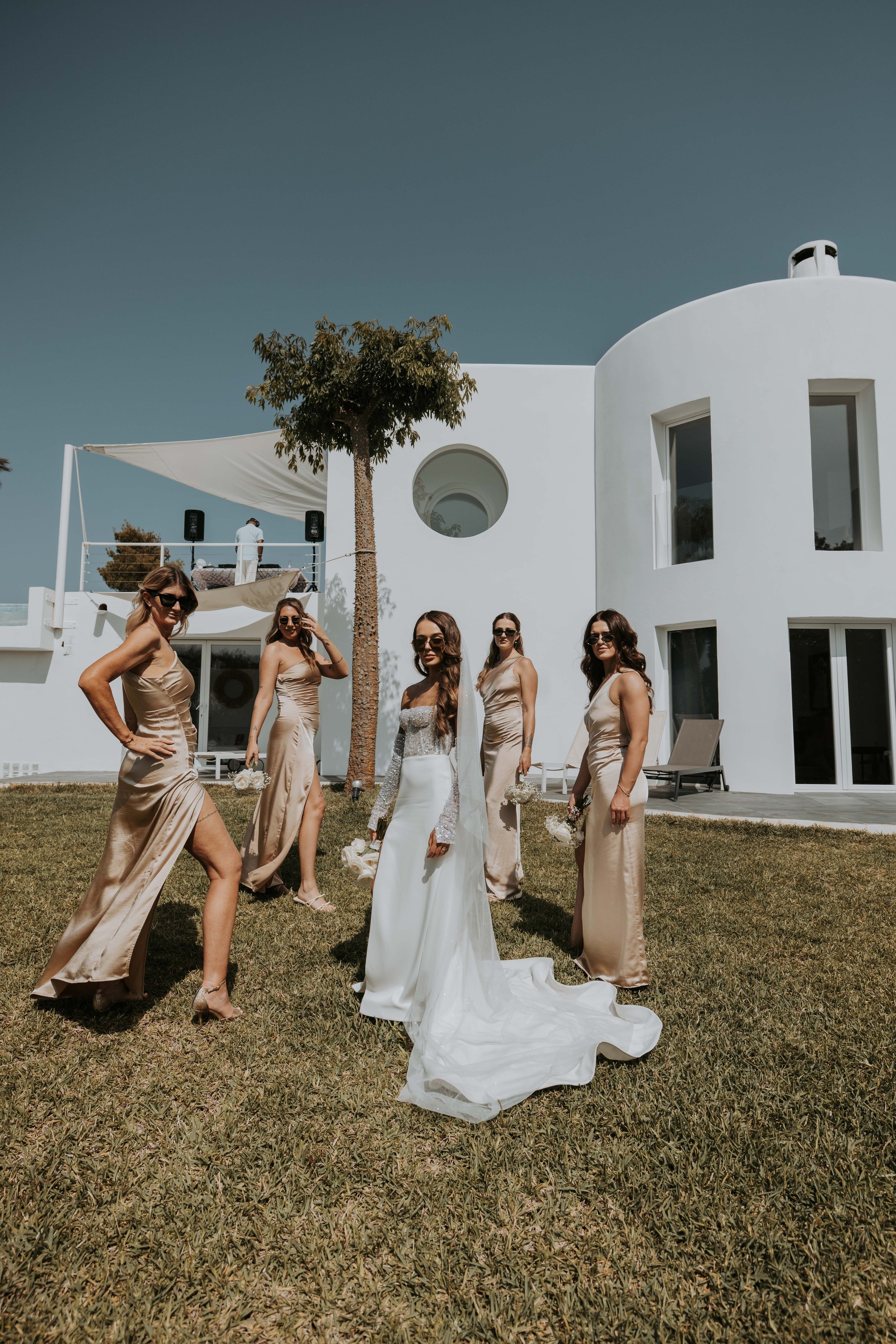 A bride in a white wedding dress surrounded by five bridesmaids in matching beige dresses, standing on a lawn in front of a modern white building with large windows and a rounded wall, under a clear blue sky.