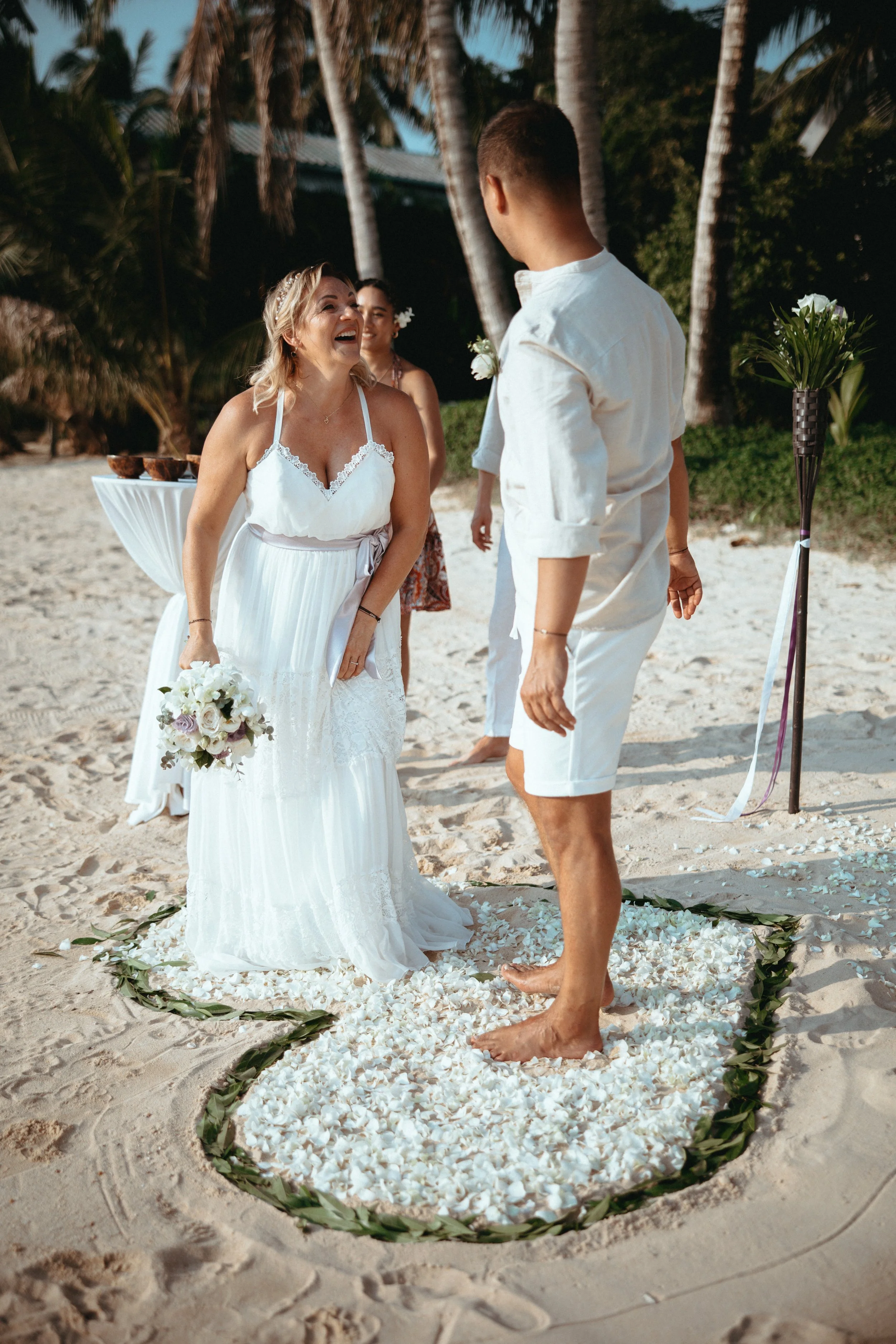 A wedding ceremony on the beach with a woman in a white dress holding flowers, a man in a white shirt and shorts, and two women in the background. They are standing on flower petals in a heart-shaped arrangement, with palm trees and sunny sky behind 