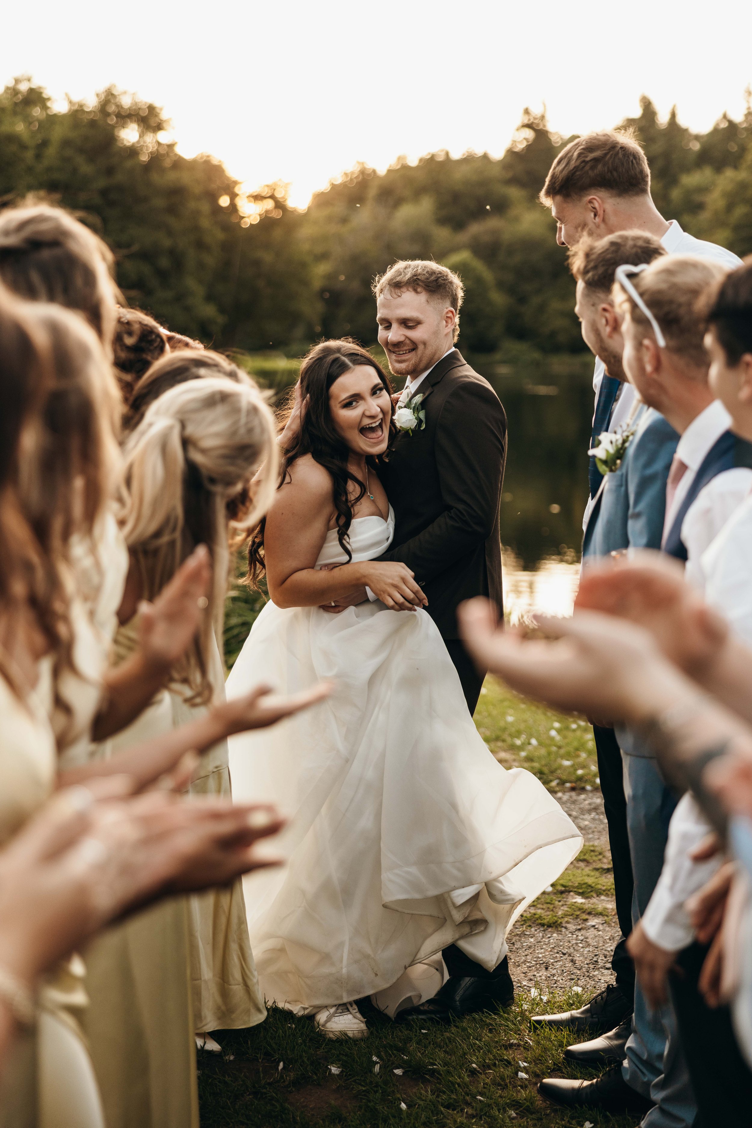 Bride and groom dancing outdoors at sunset with wedding guests clapping and smiling around them, near a lake with trees in the background.