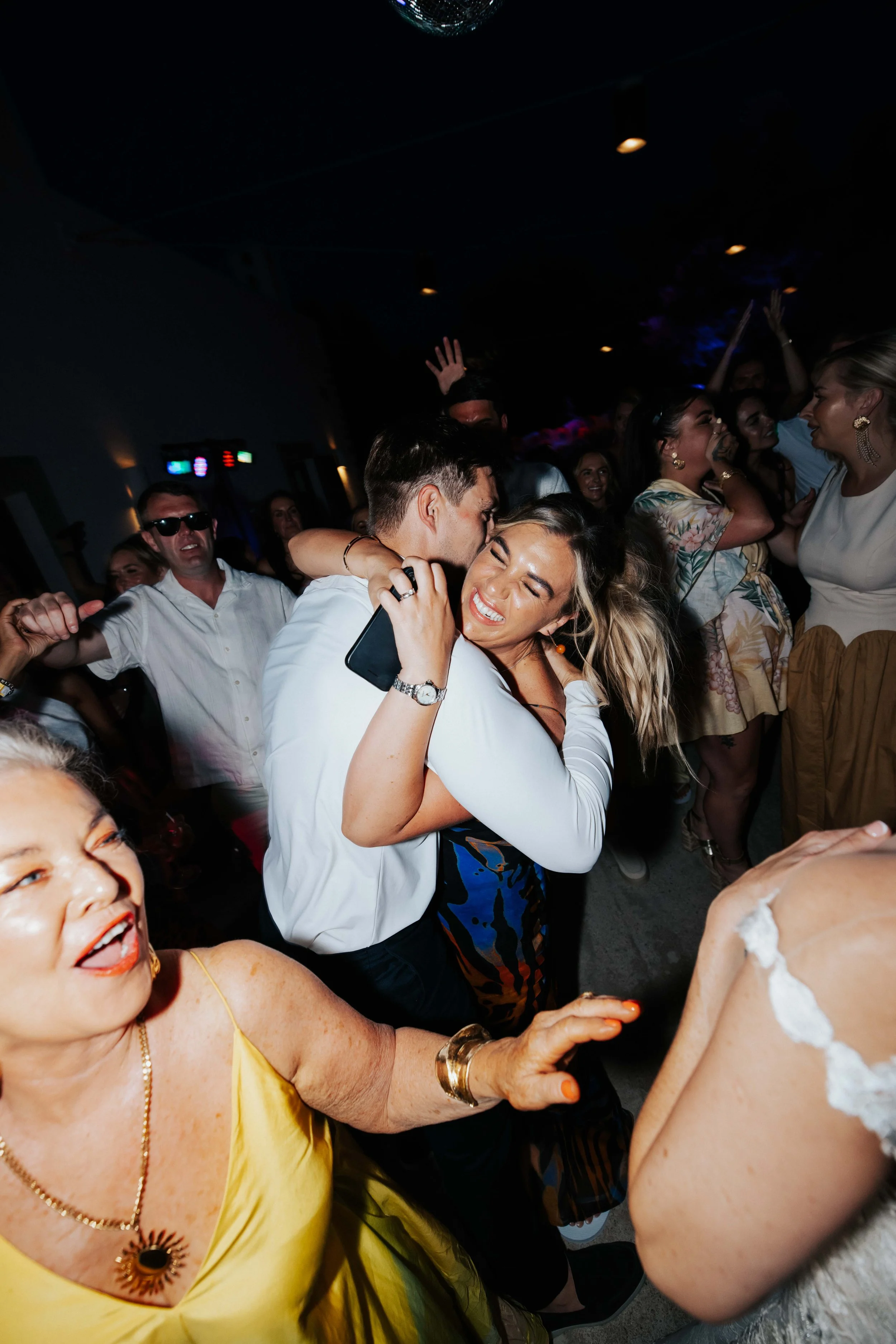 People dancing and celebrating at a party or wedding reception, with one couple hugging and others smiling and enjoying themselves.