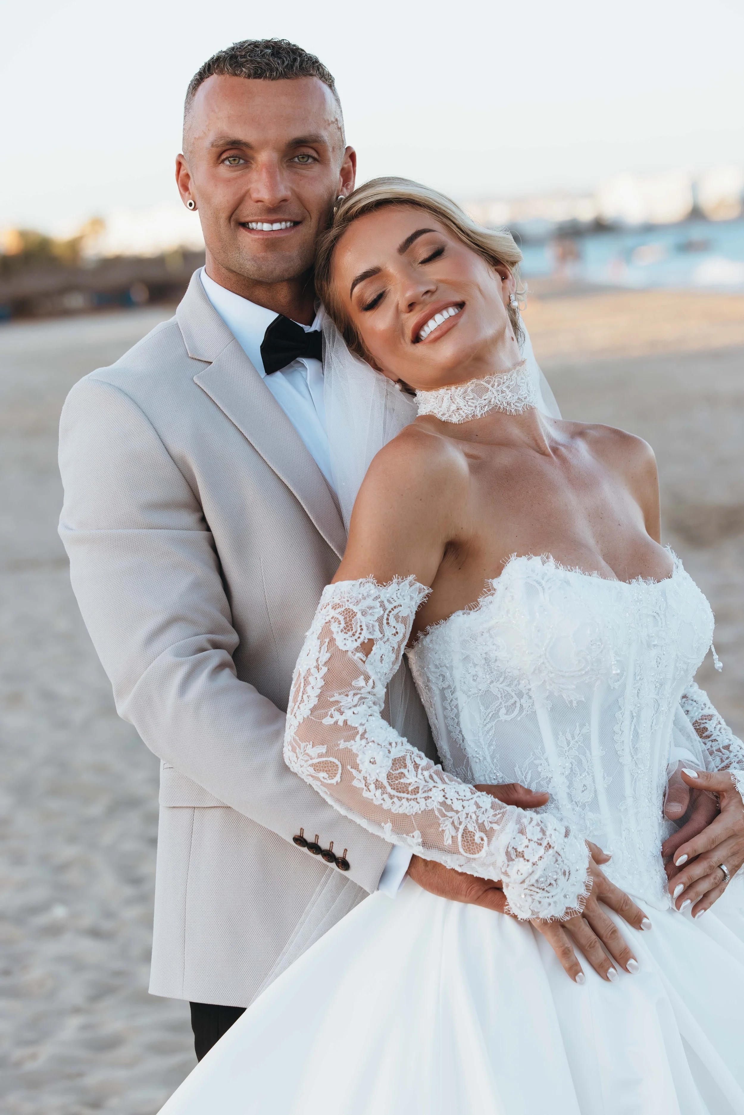A newlywed couple on the beach, with the groom in a light-colored suit and the bride in a white lace wedding gown, standing close and smiling with the ocean in the background.