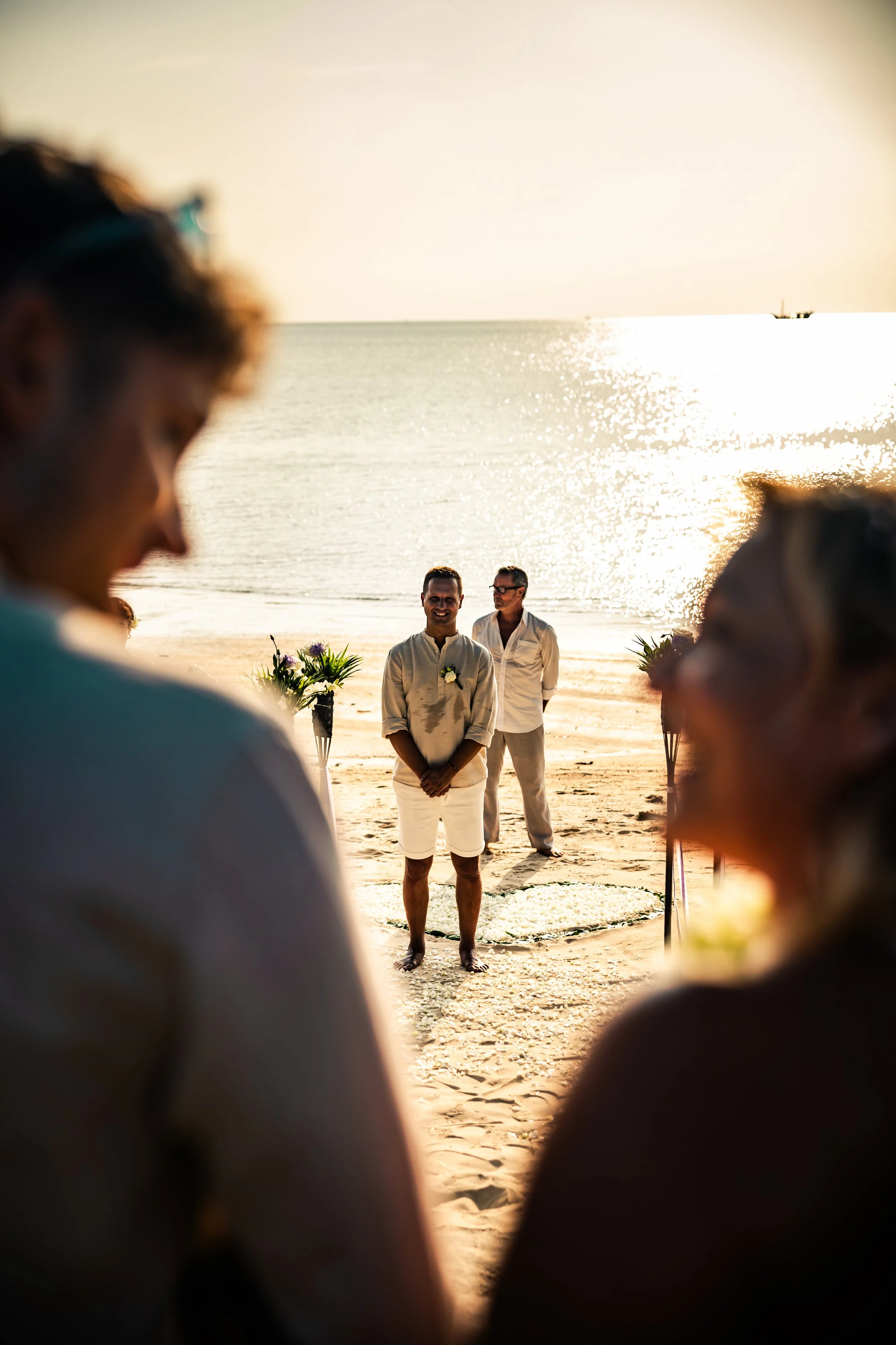 Beach wedding ceremony with two men standing in front of the ocean during sunset, seen through the blurred foreground of two people.