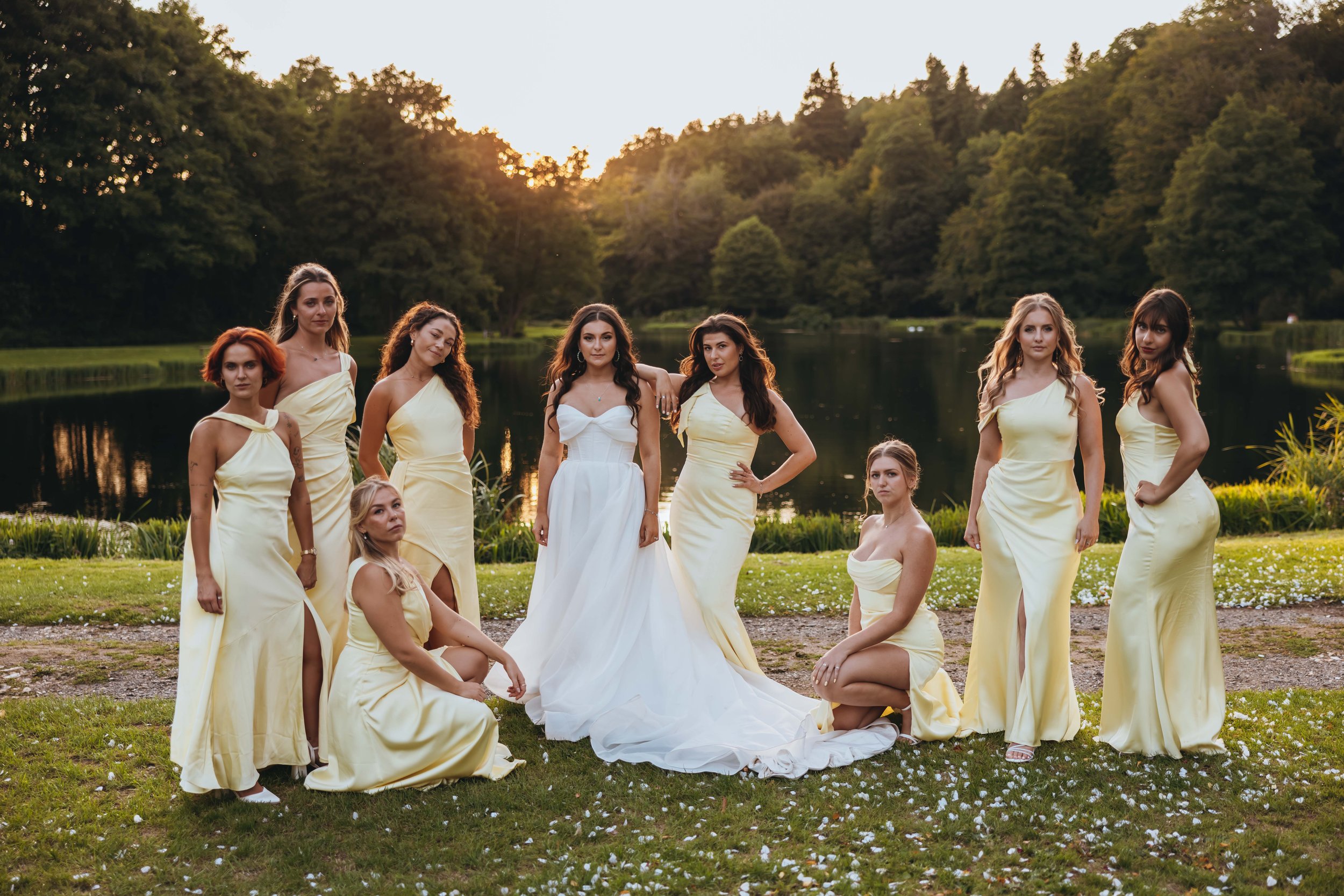 A group of ten women in evening gowns posing outdoors near a lake at sunset, with trees in the background.