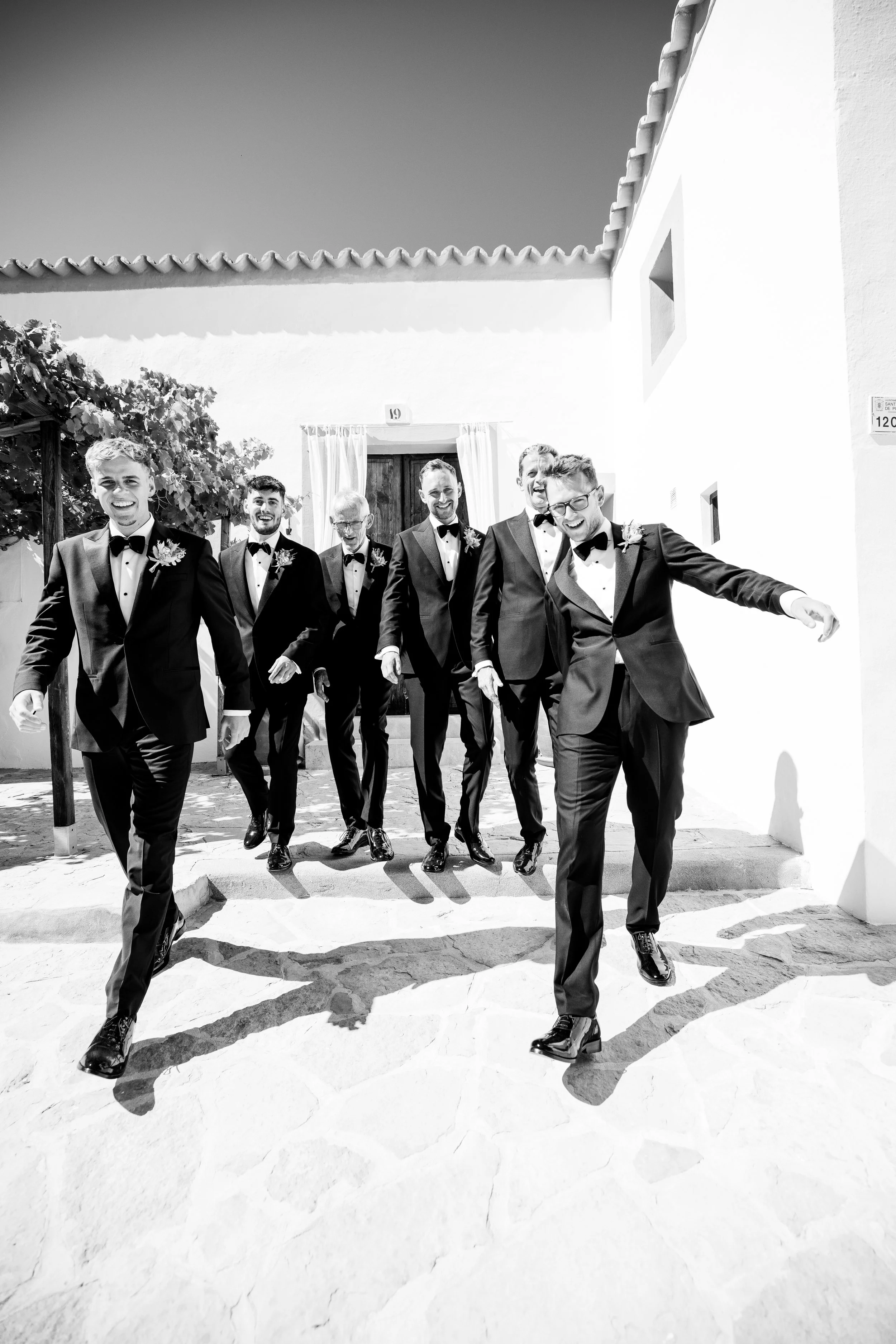 A group of six men in tuxedos walking outdoors, smiling and laughing, with a white building in the background.