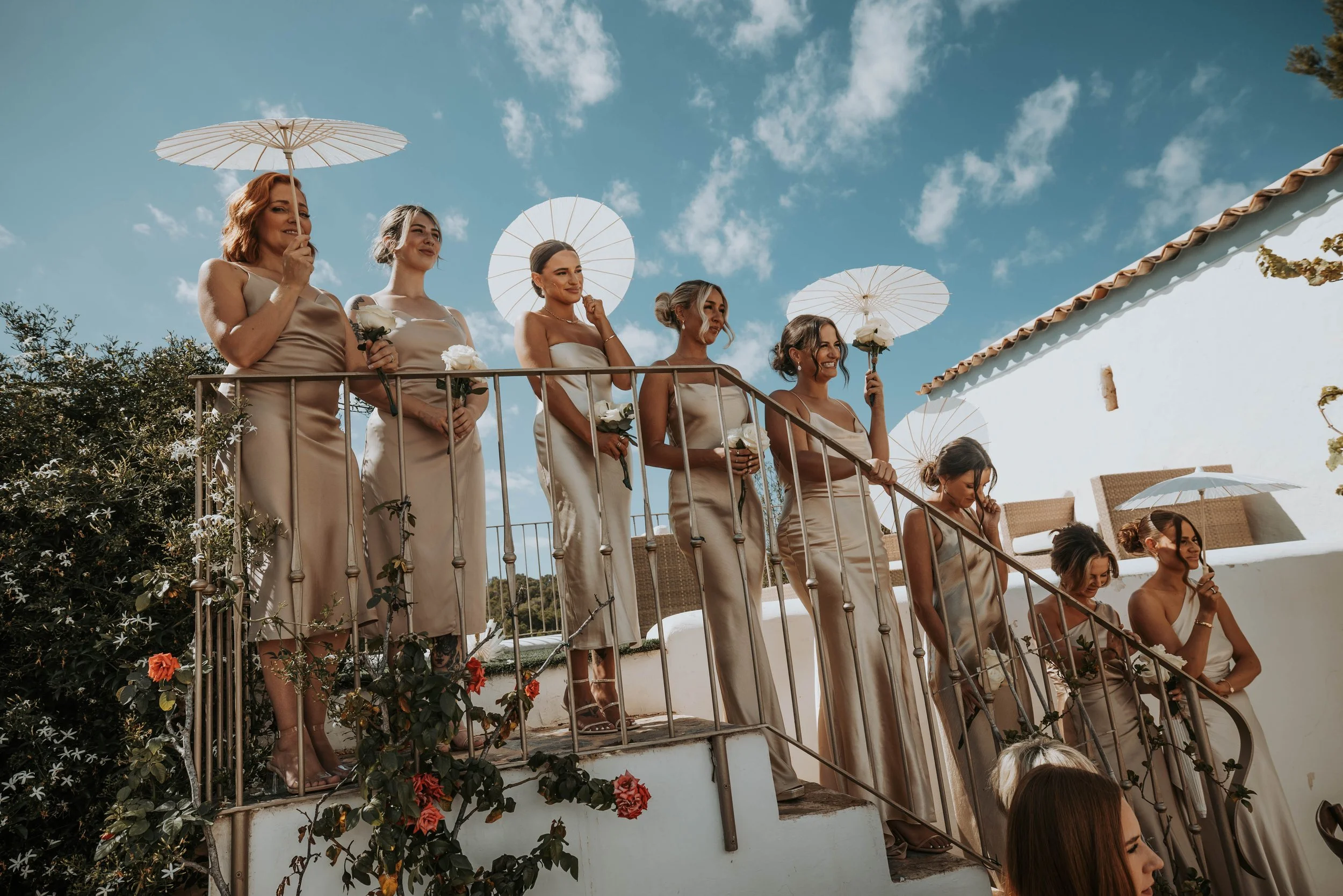A group of women dressed in beige dresses are standing on a staircase outdoors, holding umbrellas and white roses, during a wedding ceremony under a blue sky with scattered clouds.