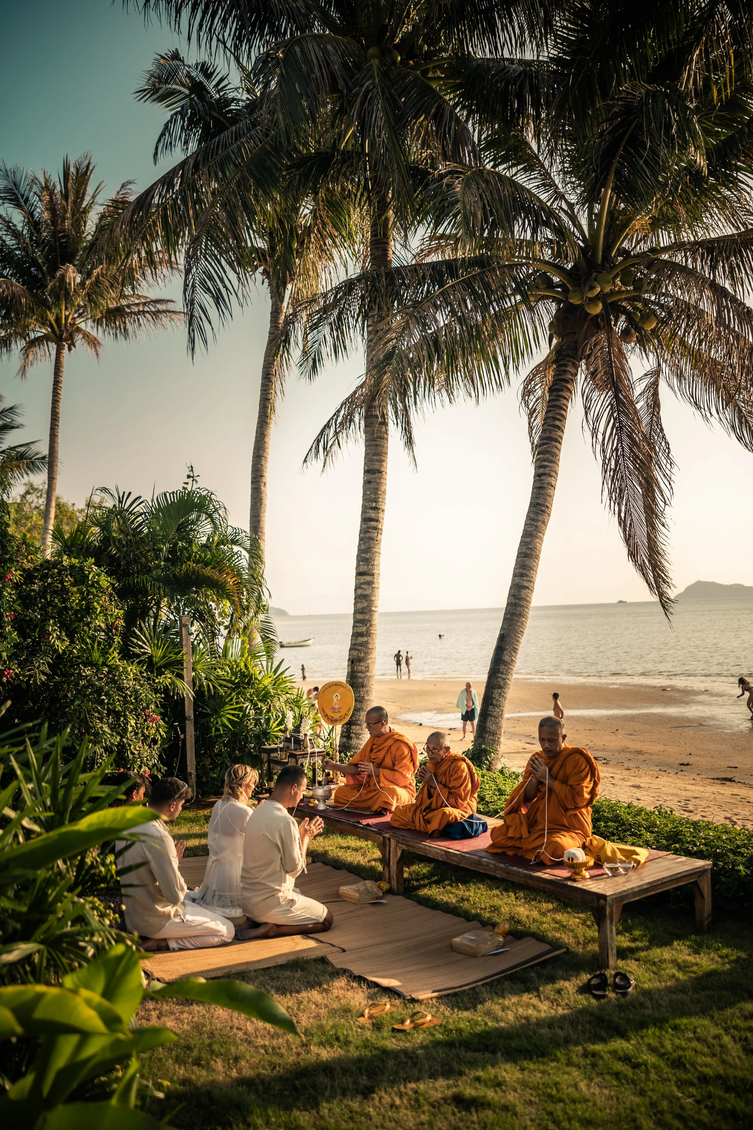 People engaged in a religious ceremony on a grassy area near a beach, with palm trees and the ocean in the background during sunset.