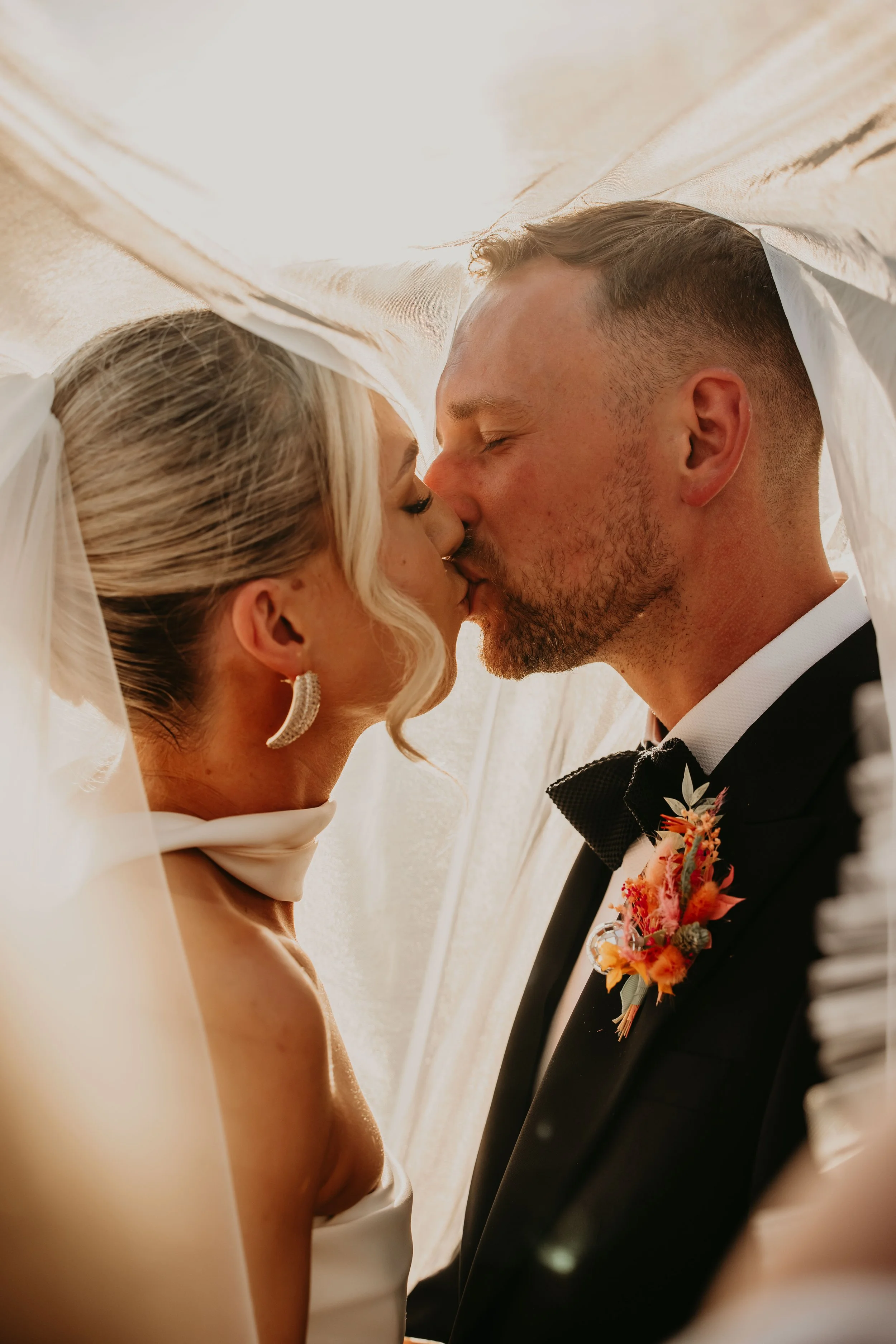 A bride and groom share a kiss inside a wedding veil with soft lighting.
