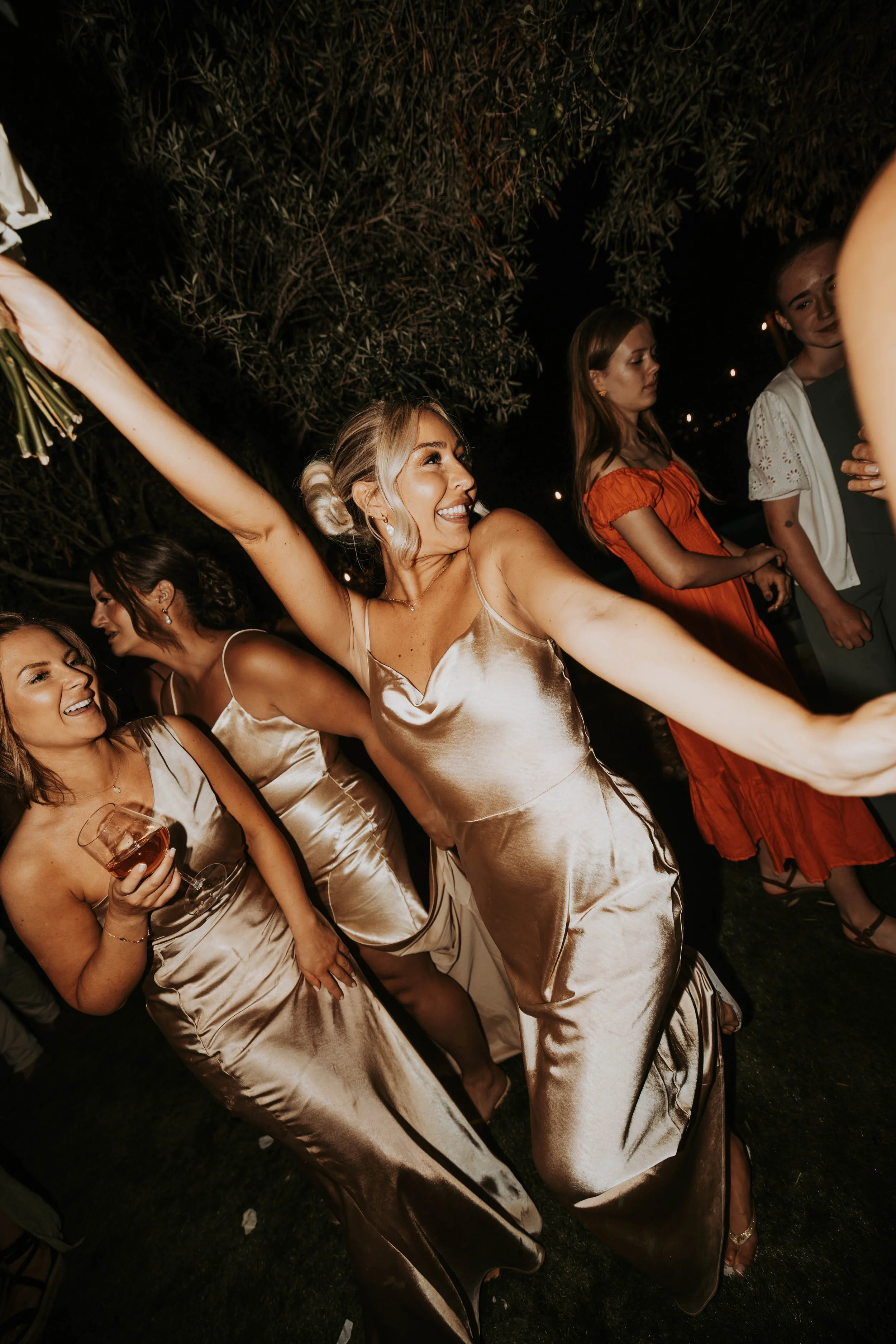 Group of women celebrating at a nighttime outdoor event, wearing elegant satin dresses, smiling, and holding drinks.
