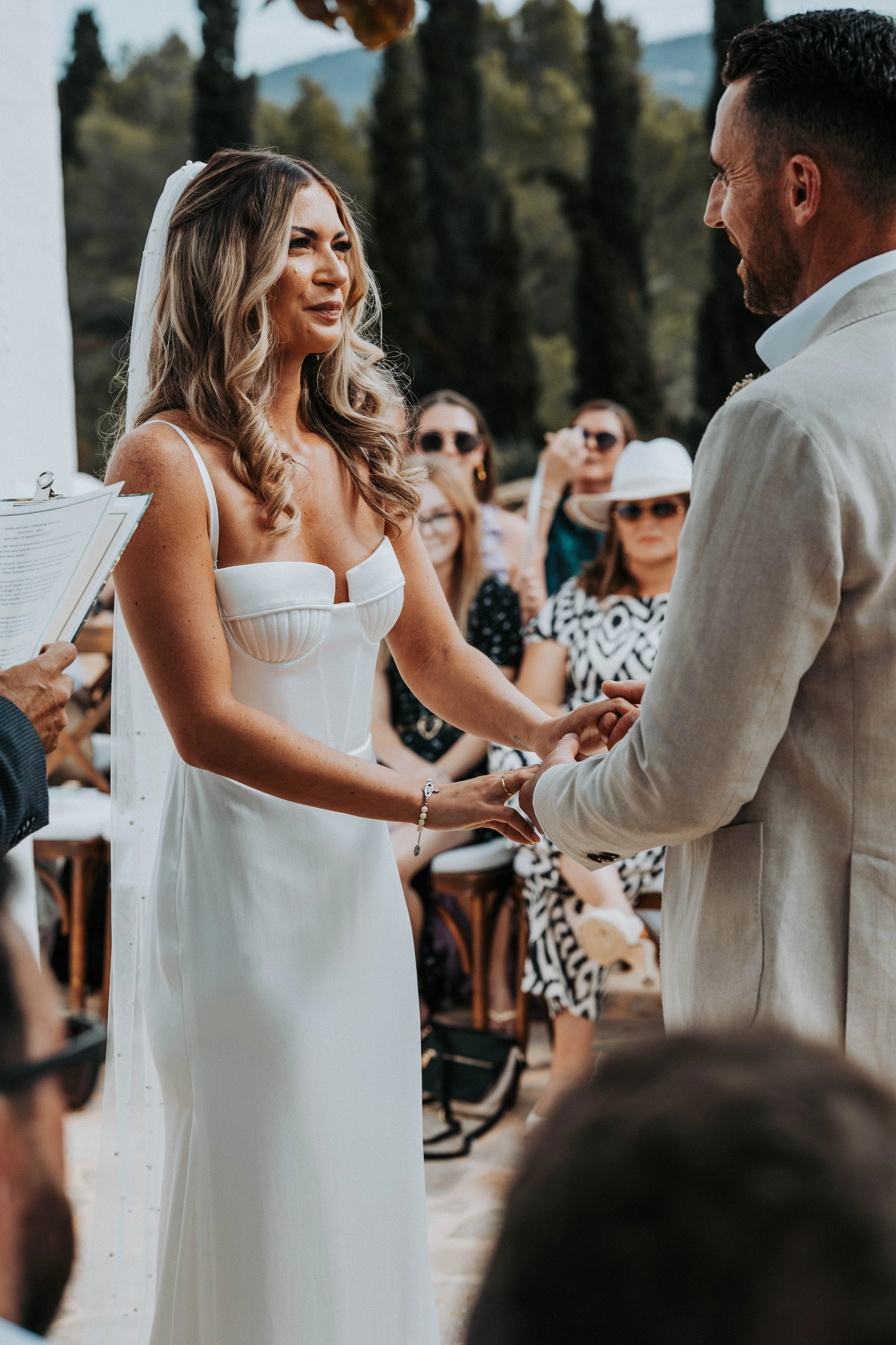 A bride and groom exchange vows during an outdoor wedding ceremony, with guests seated in the background and trees and mountains in the distance.