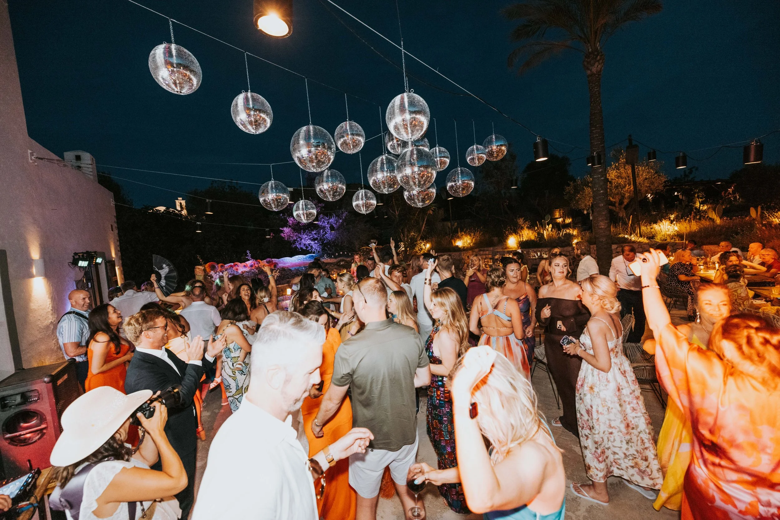 People dancing and socializing at an outdoor party during the evening with disco balls hanging overhead and palm trees in the background.