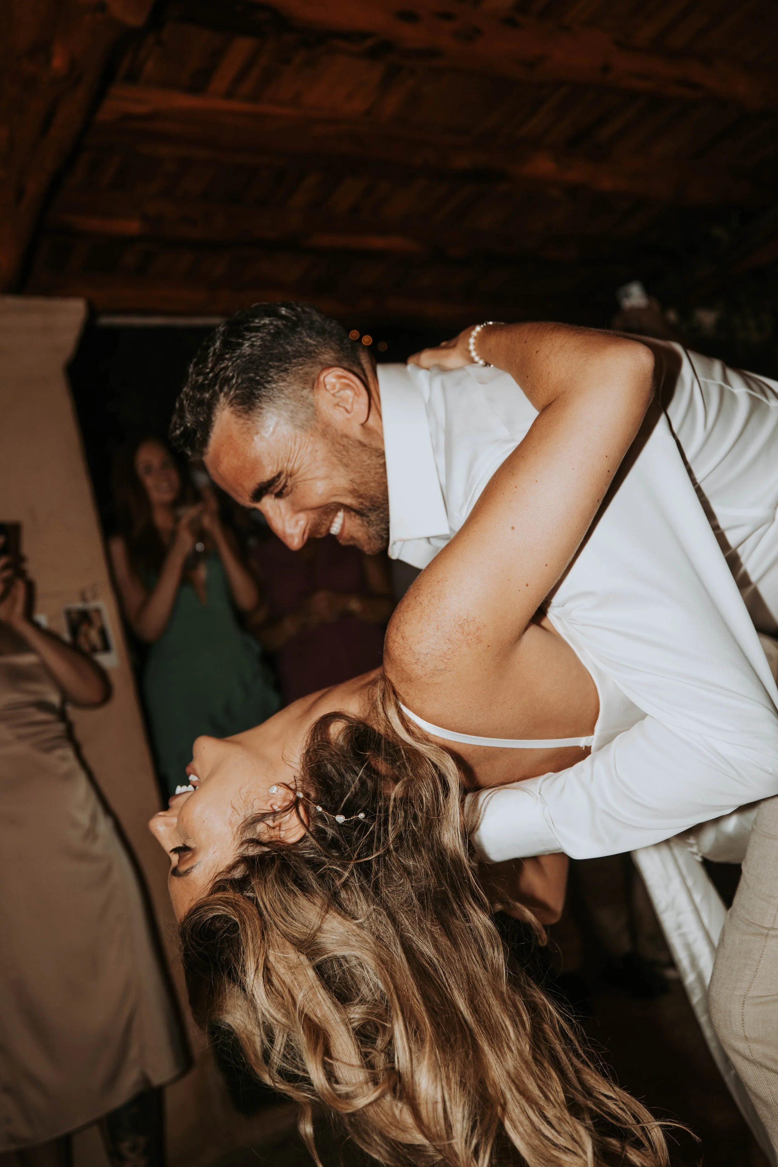 A couple sharing a dance, with the man leaning down and smiling while the woman leans back and laughs. They are at a celebration or wedding reception, with guests in the background taking photos.
