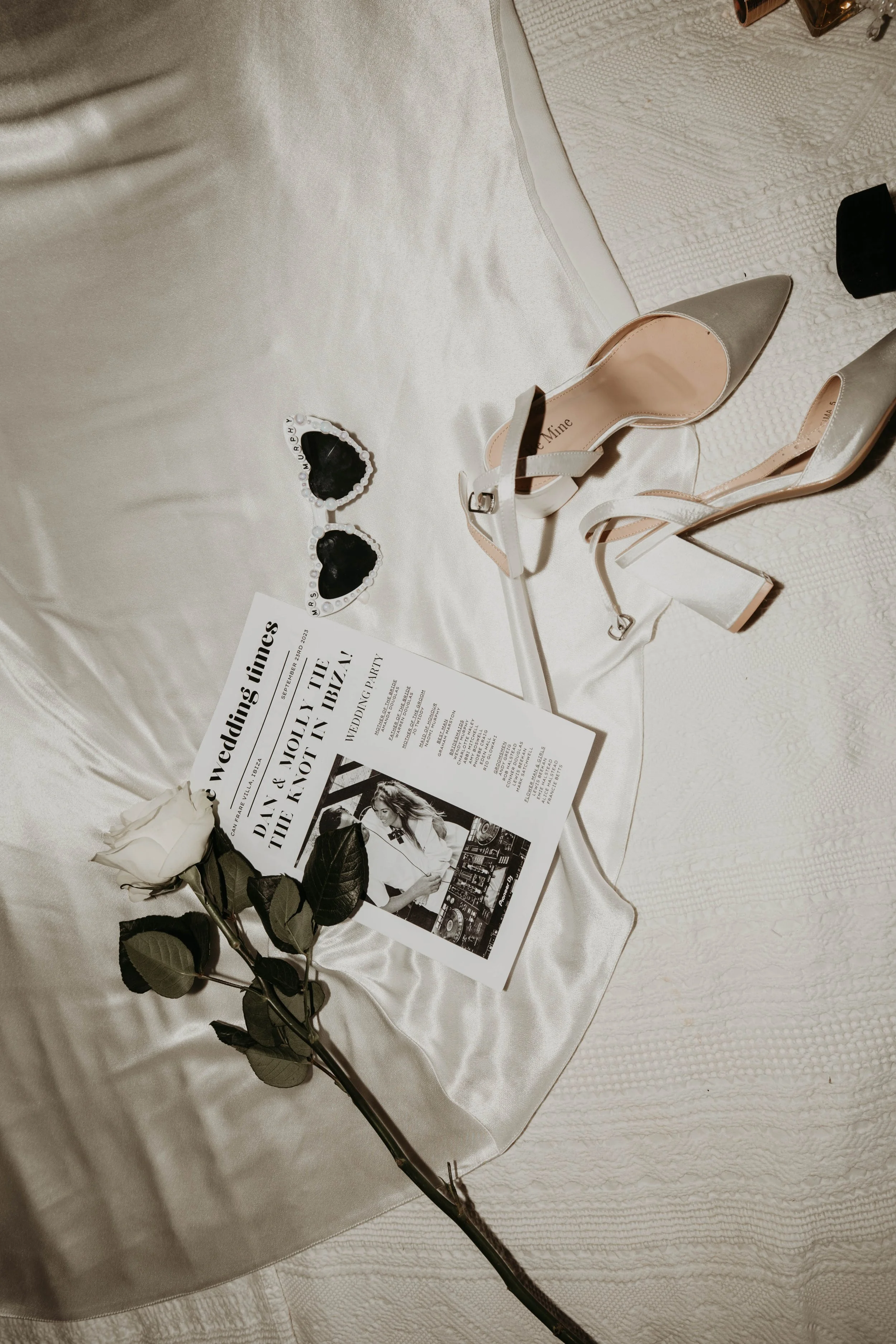 A wedding invitation, a pair of beige high-heeled shoes, a pair of heart-shaped sunglasses, and a white rose lay on a satin cloth.