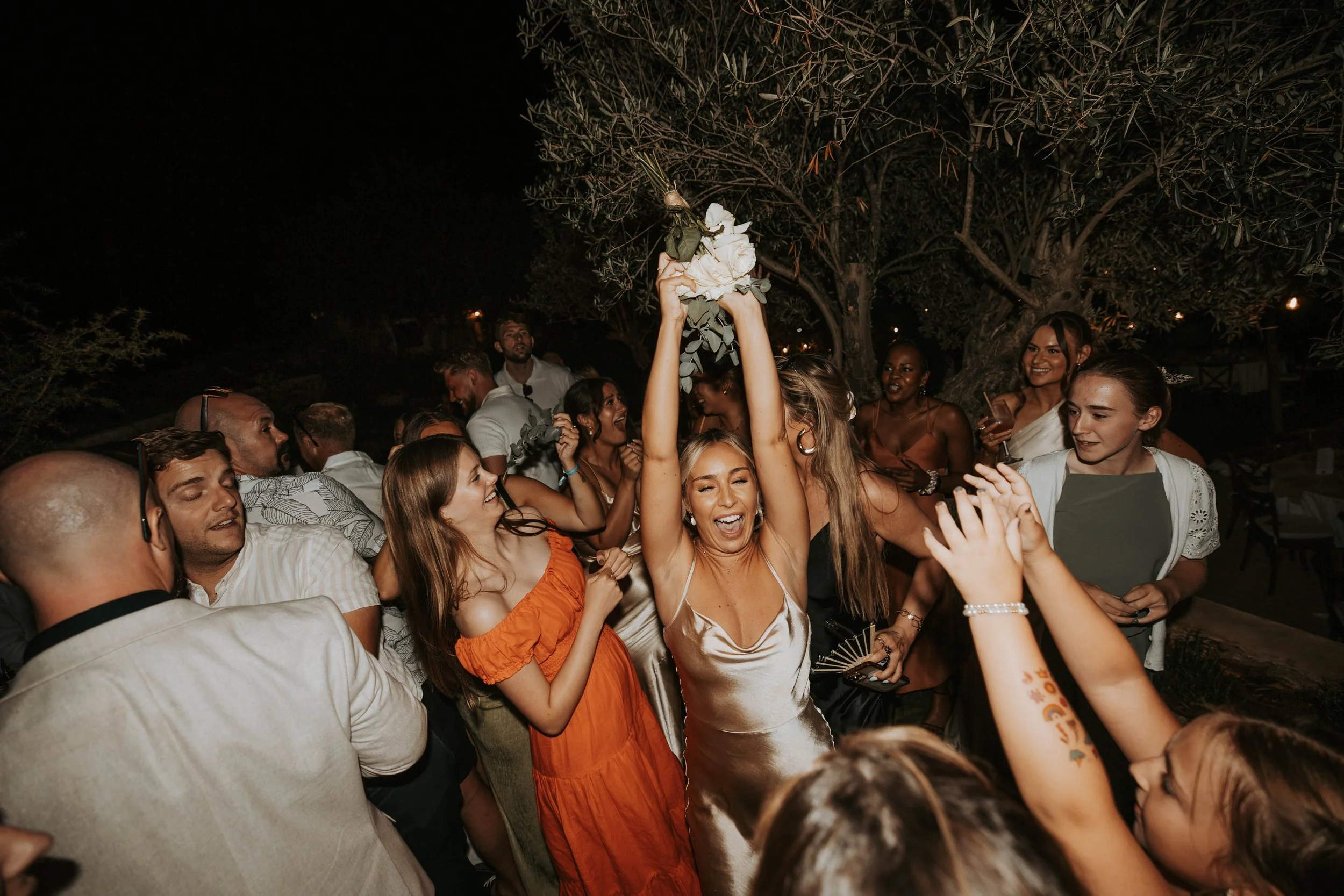 A woman in a champagne-colored dress holding a bouquet above her head, surrounded by a joyful crowd celebrating outdoors at night.