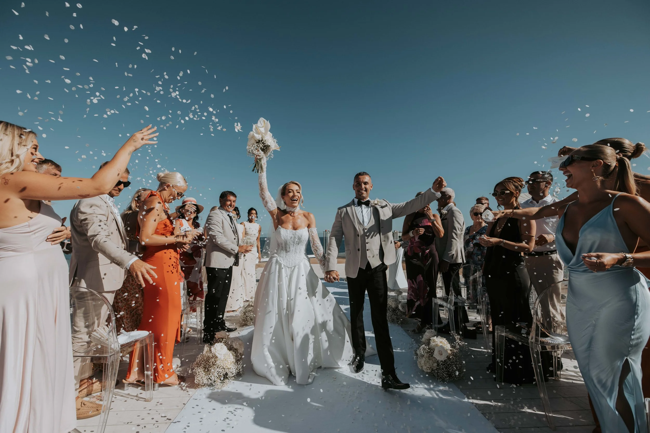 A bride and groom walk hand in hand down an outdoor aisle, smiling, as friends and family shower them with confetti on a sunny day.