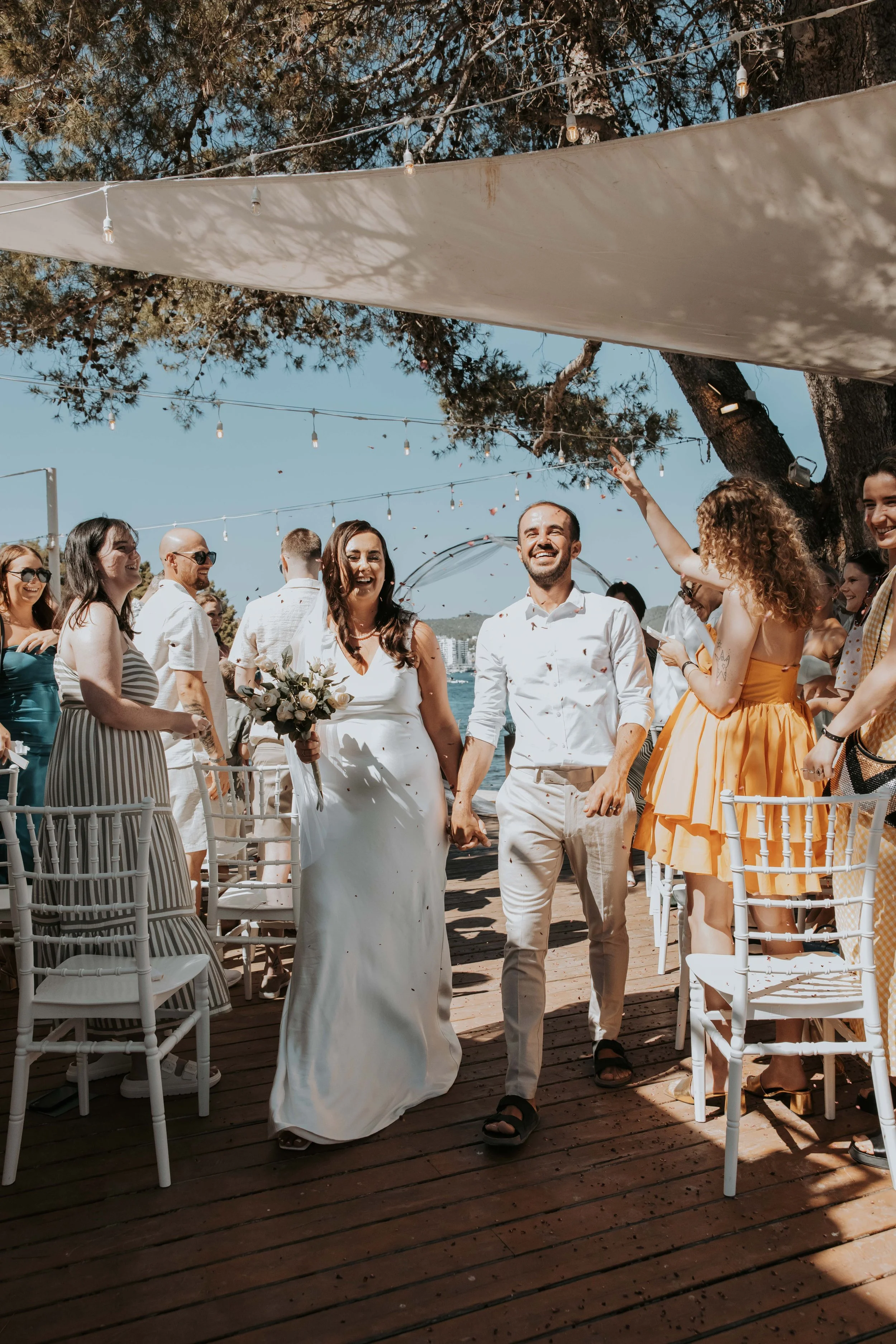 A couple in wedding attire walking hand-in-hand down an outdoor wooden aisle, smiling and celebrating. They are surrounded by guests, some seated and some standing, with a large tree, string lights, and blue sky in the background.