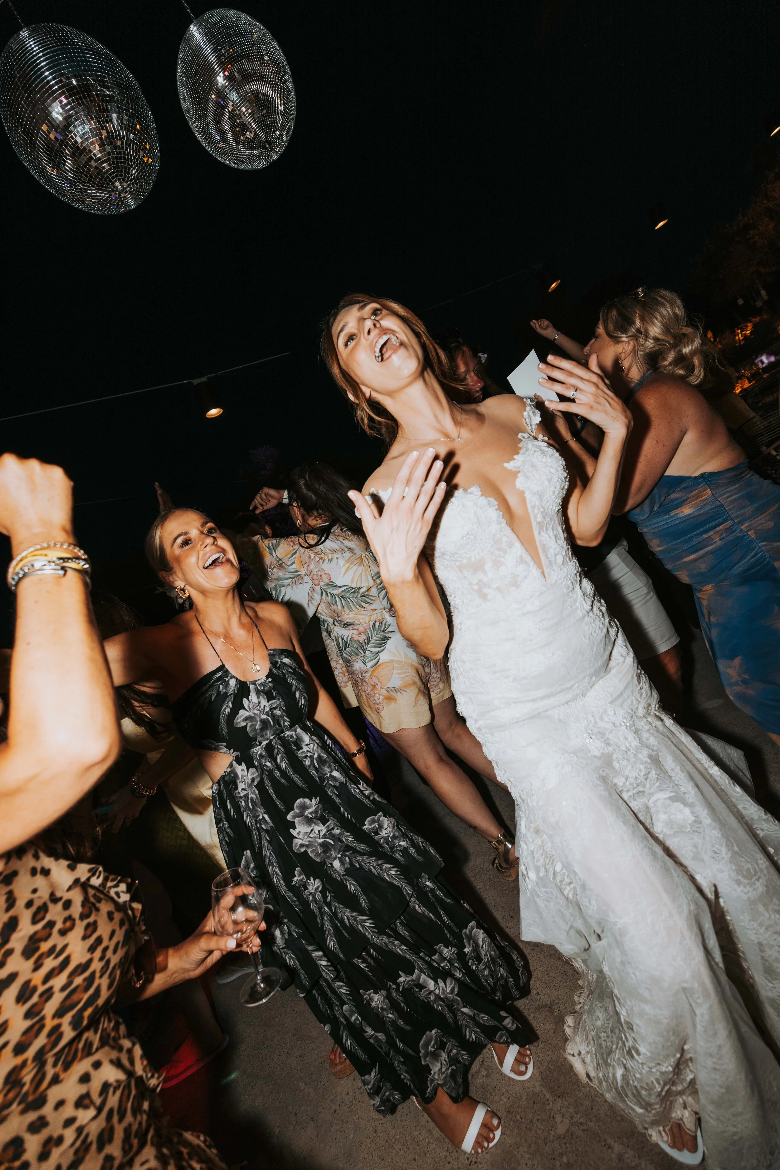 A bride in a white wedding dress dancing and celebrating with friends at a nighttime wedding reception, with disco balls overhead.