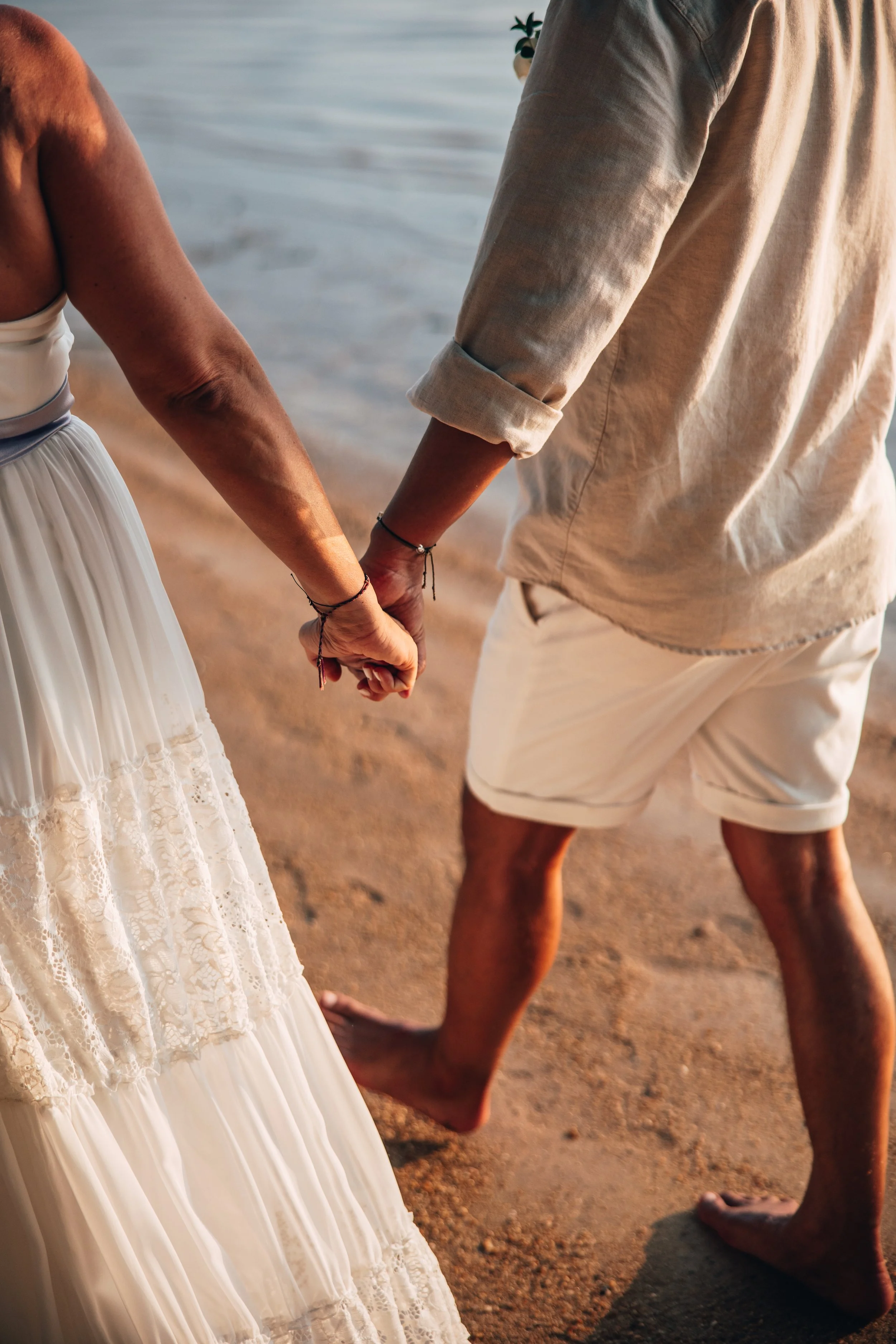 A couple holding hands on the beach, with the woman wearing a white lace dress and the man wearing a beige shirt and shorts.
