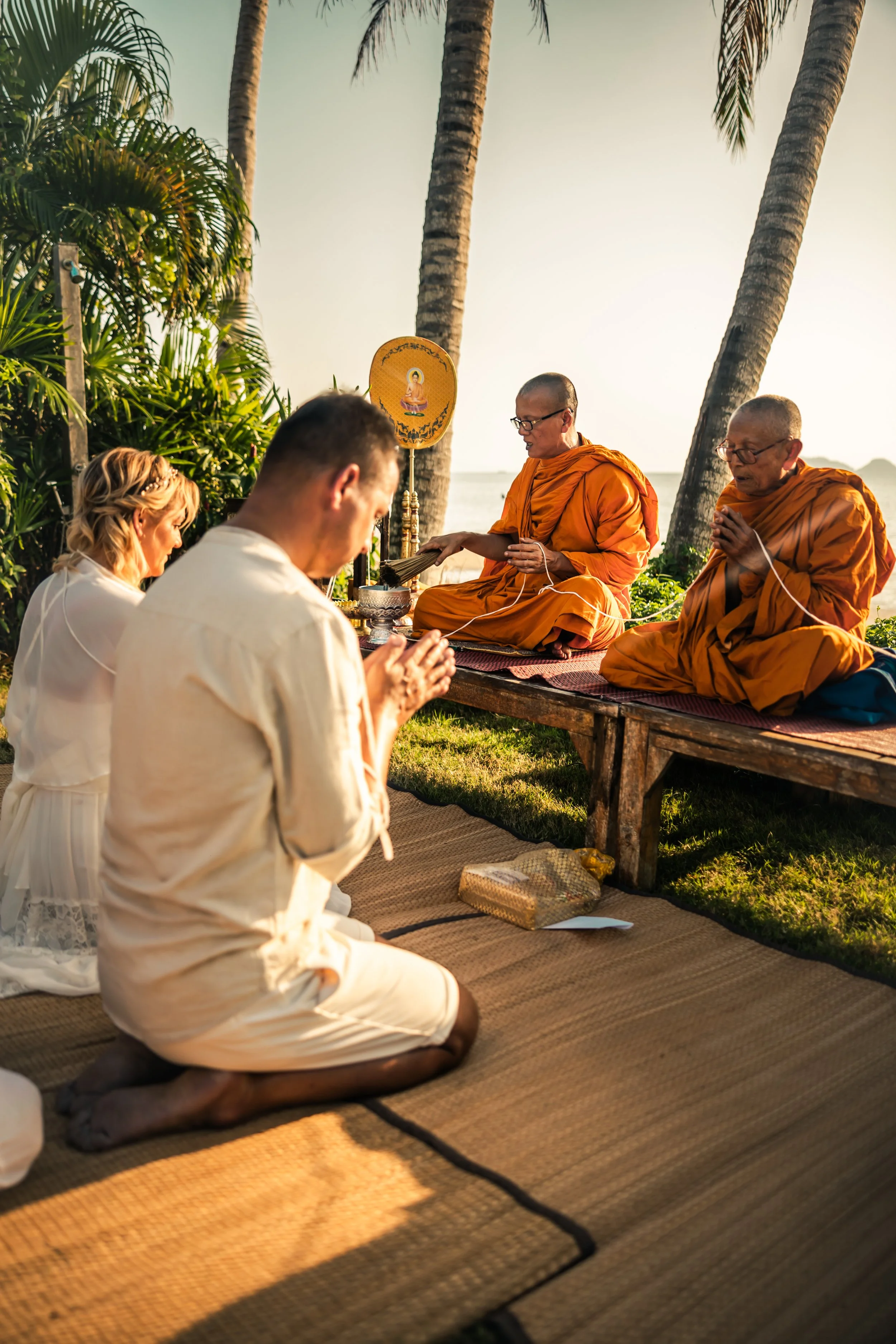 A couple is praying outdoors on a rug, facing two Buddhist monks dressed in orange robes, with a scenic background of palm trees, lush greenery, and a sunset over the ocean.
