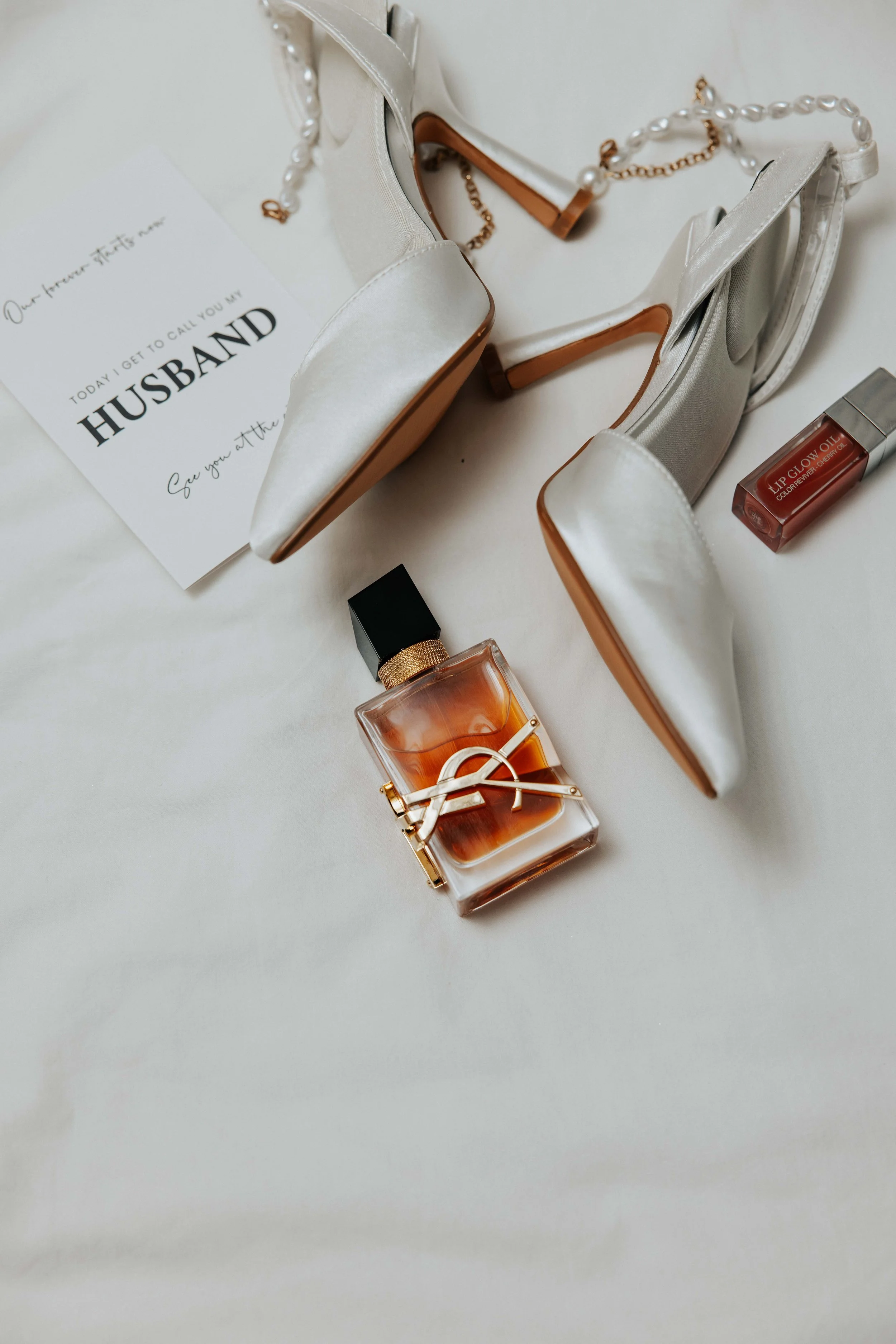 A white silk high-heeled shoe with a chain and pearl strap, a bottle of YSL perfume, lip glow oil, and a note that reads, 'Our forever starts one today! I get to call you my husband. See you at the...
