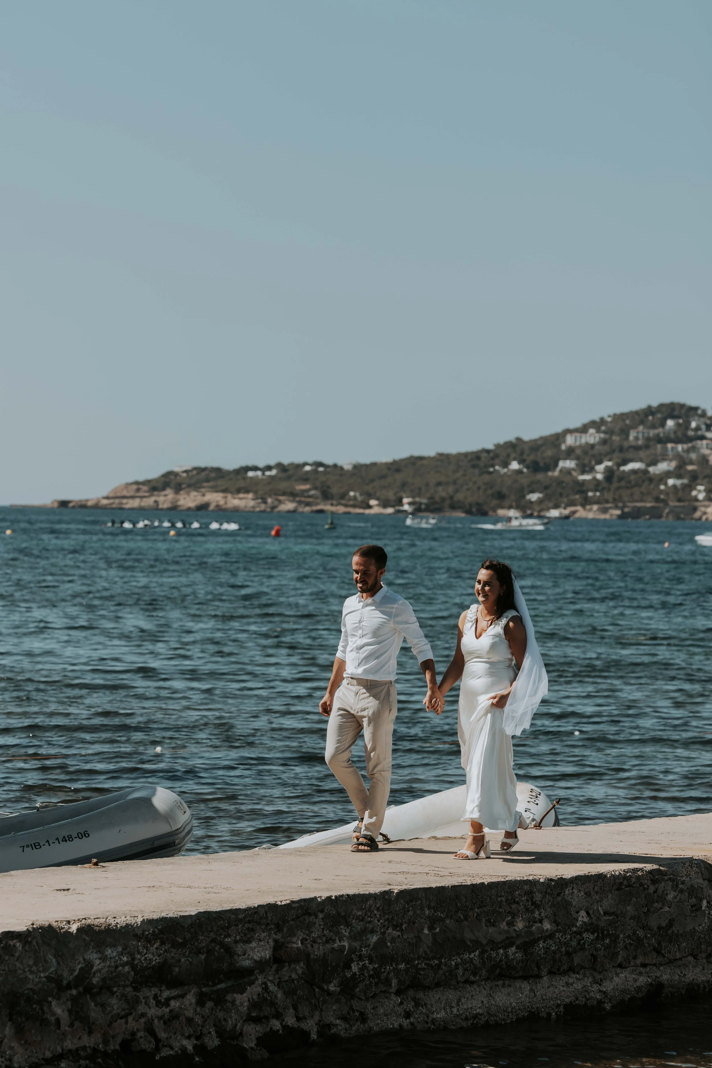 A couple in wedding attire walking along a beachside pier, holding hands, with the ocean and distant shoreline in the background.