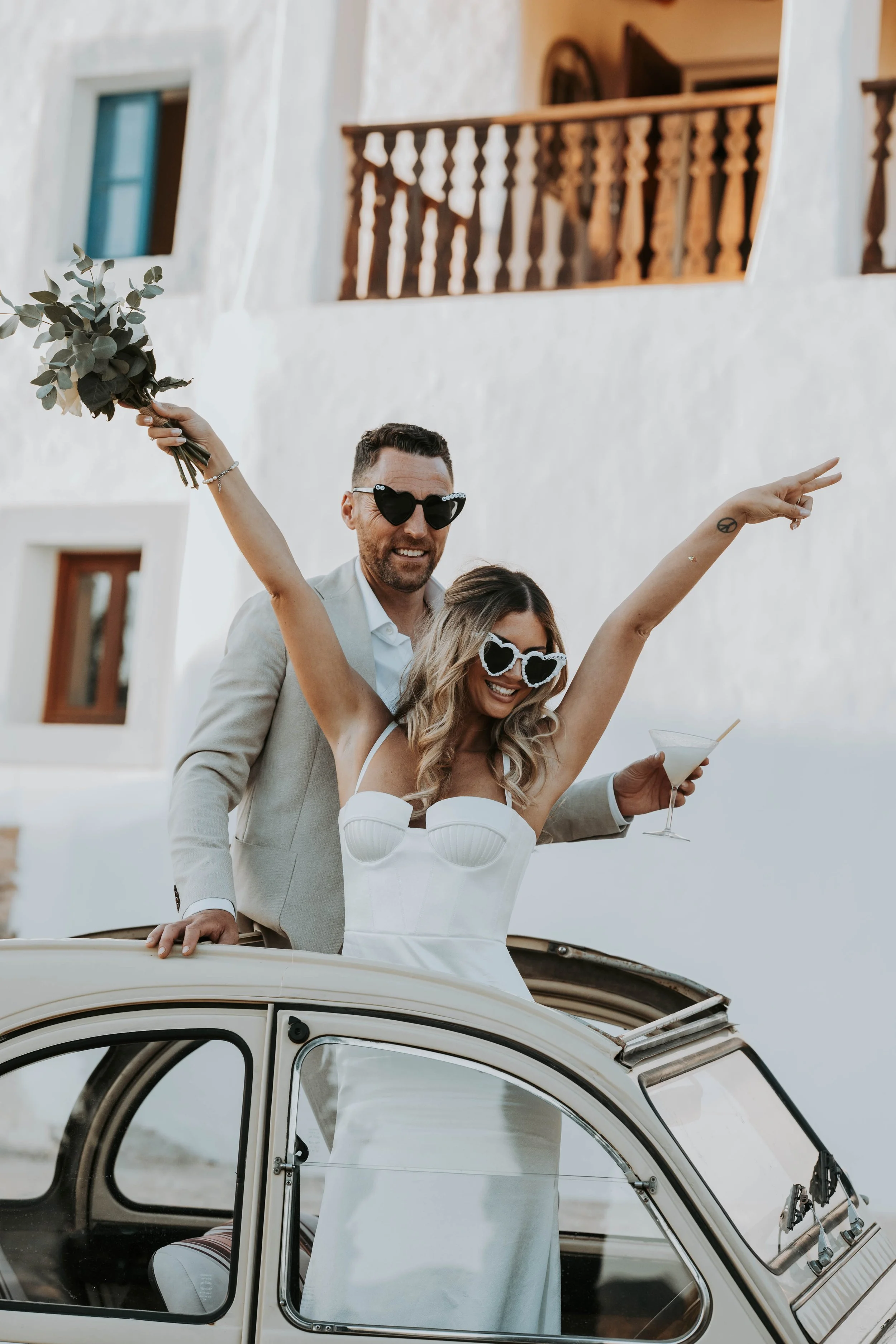 A newlywed couple celebrating from the sunroof of a classic car, with the woman wearing heart-shaped sunglasses and a white wedding dress, holding a cocktail, and the man in a suit with sunglasses, holding a bouquet.