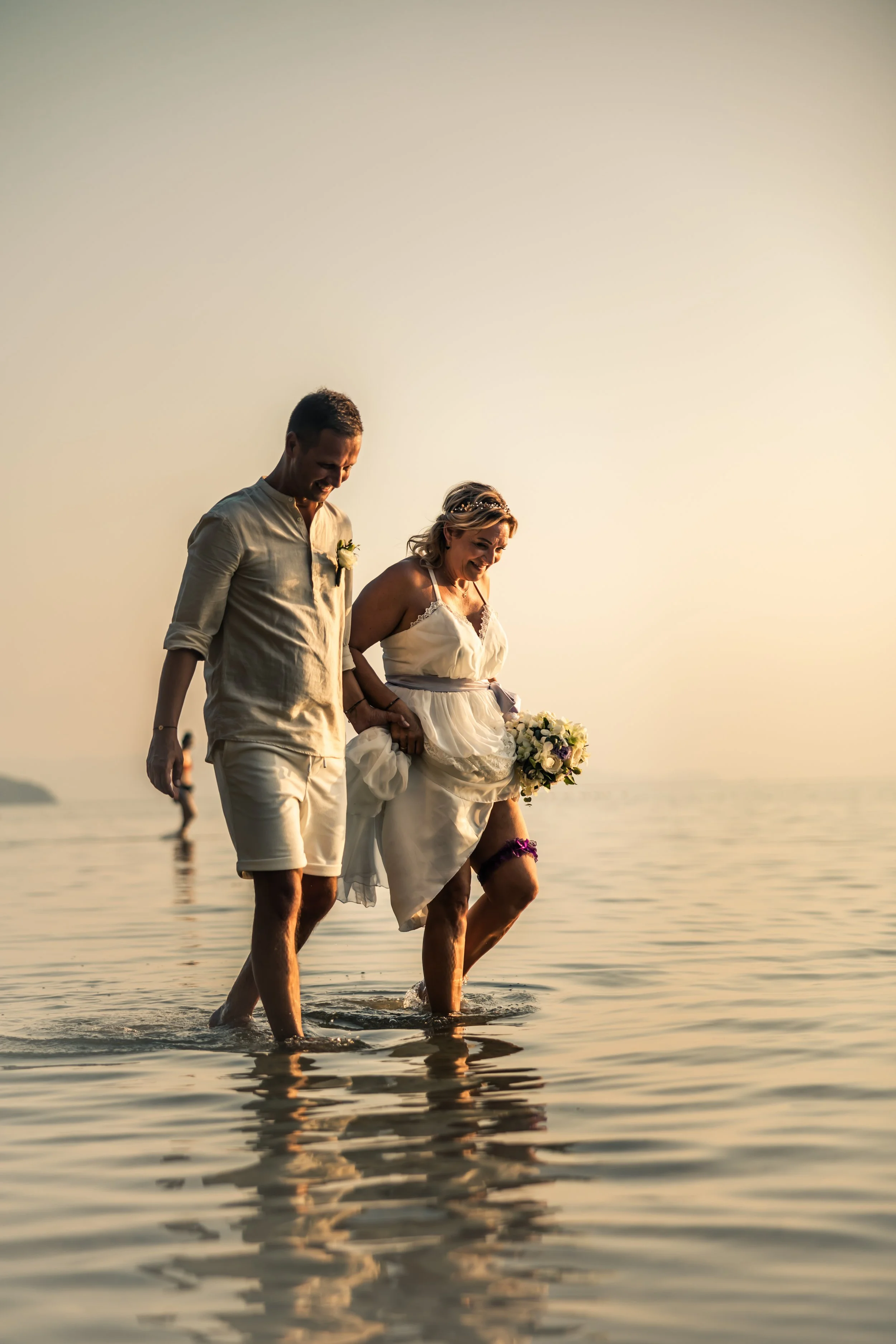 A couple in wedding attire walking in shallow water at sunset, holding a bouquet, with a relaxed and joyful expression.
