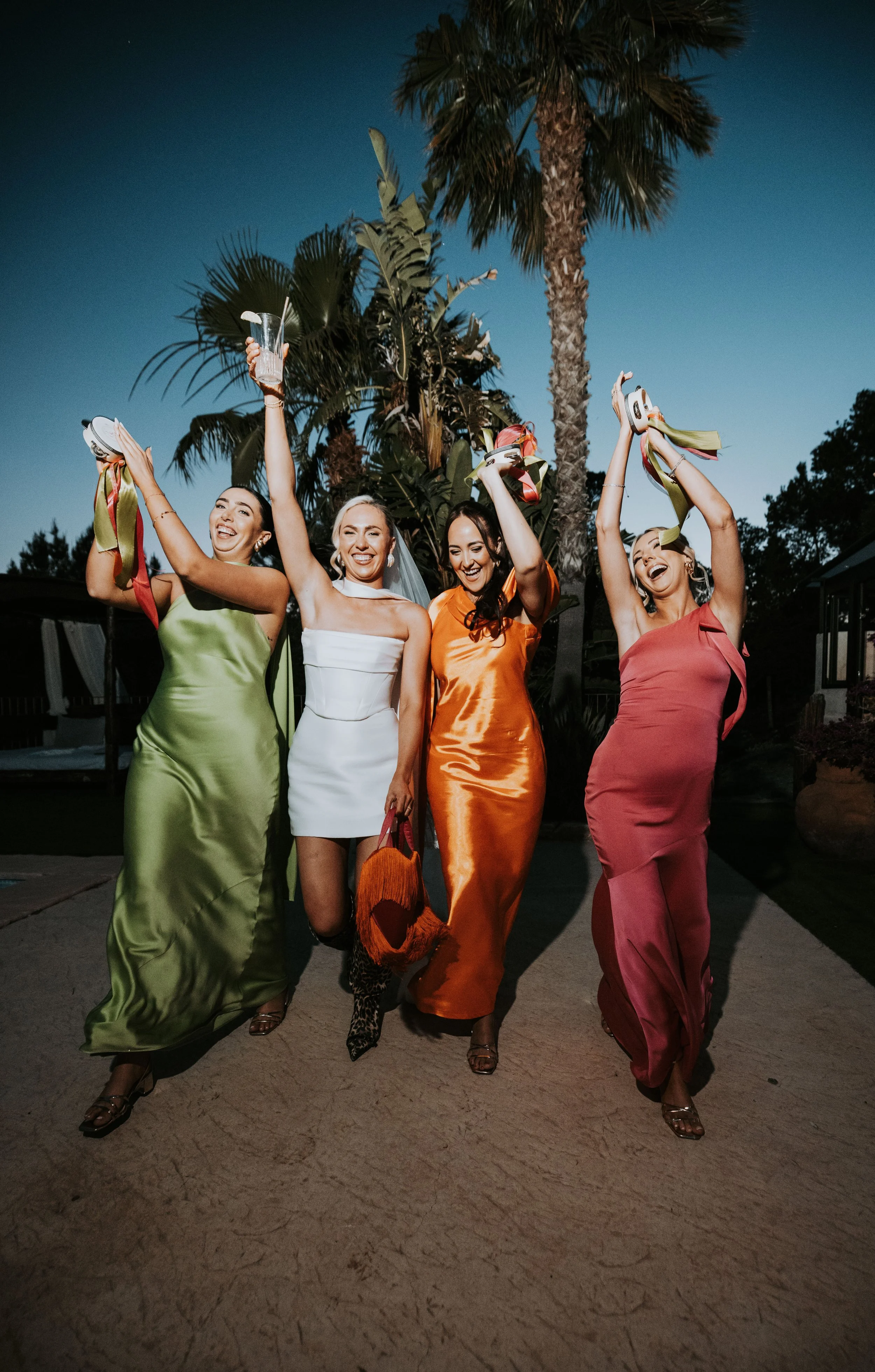 Four women in colorful satin dresses celebrating outdoors at dusk, holding up shoes and a drink, smiling and walking on a pathway with palm trees in the background.