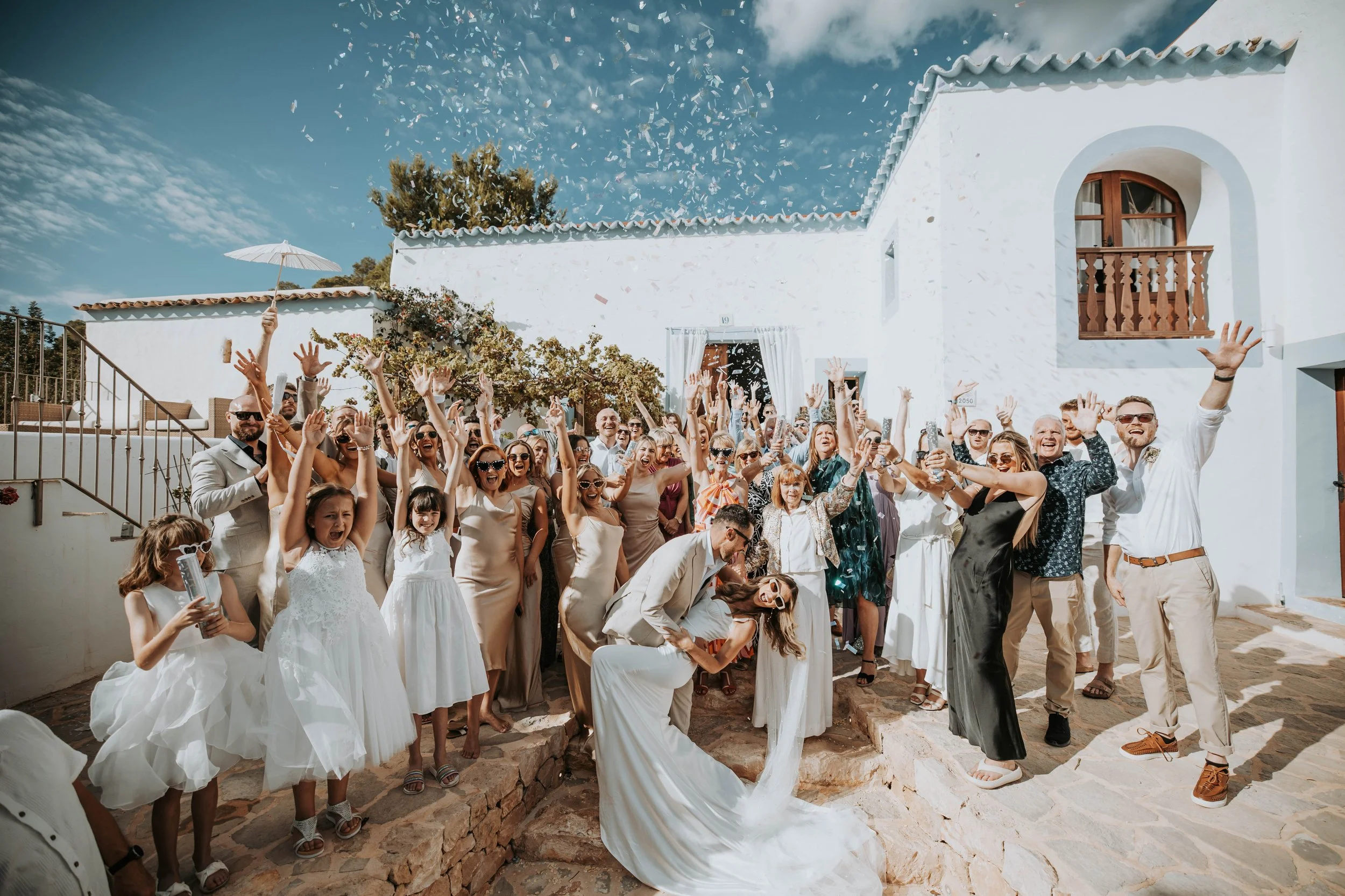 A large group of people celebrating outdoors, with a bride and groom in the center kissing while others cheer and throw confetti. The setting is sunny with a white building in the background.
