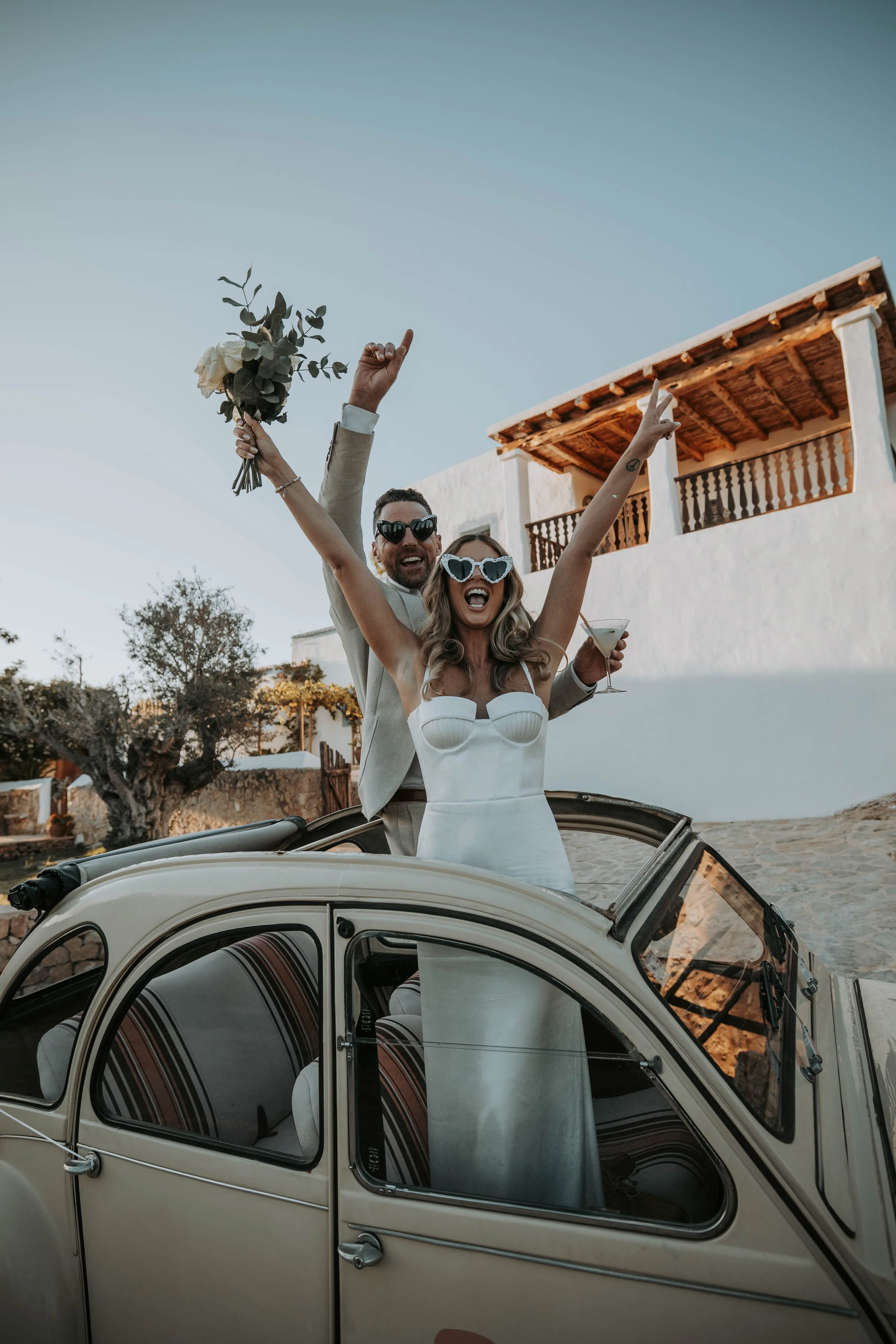 A couple in wedding attire celebrating on a vintage car, with the woman in a white dress and heart-shaped sunglasses, holding a bouquet and a cocktail, while the man in a beige suit and sunglasses stands behind her, raising their hands in joy.