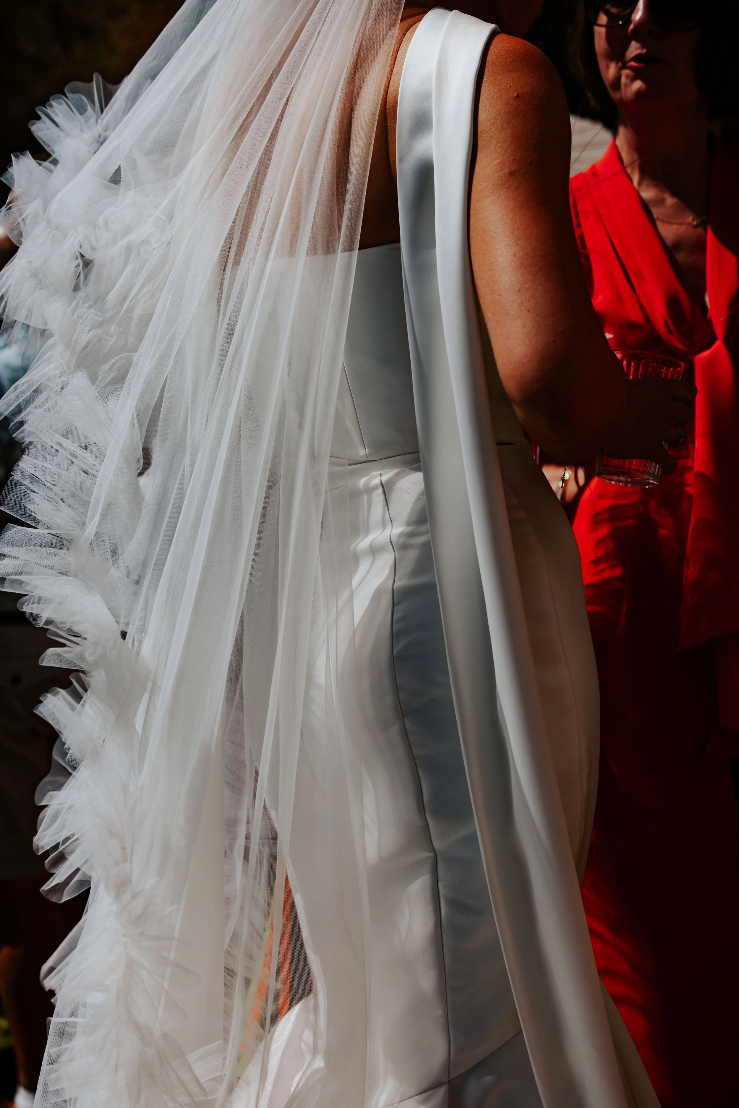 Close-up of a bride in a white wedding gown with a long, flowy veil, talking to a woman in a red outfit.