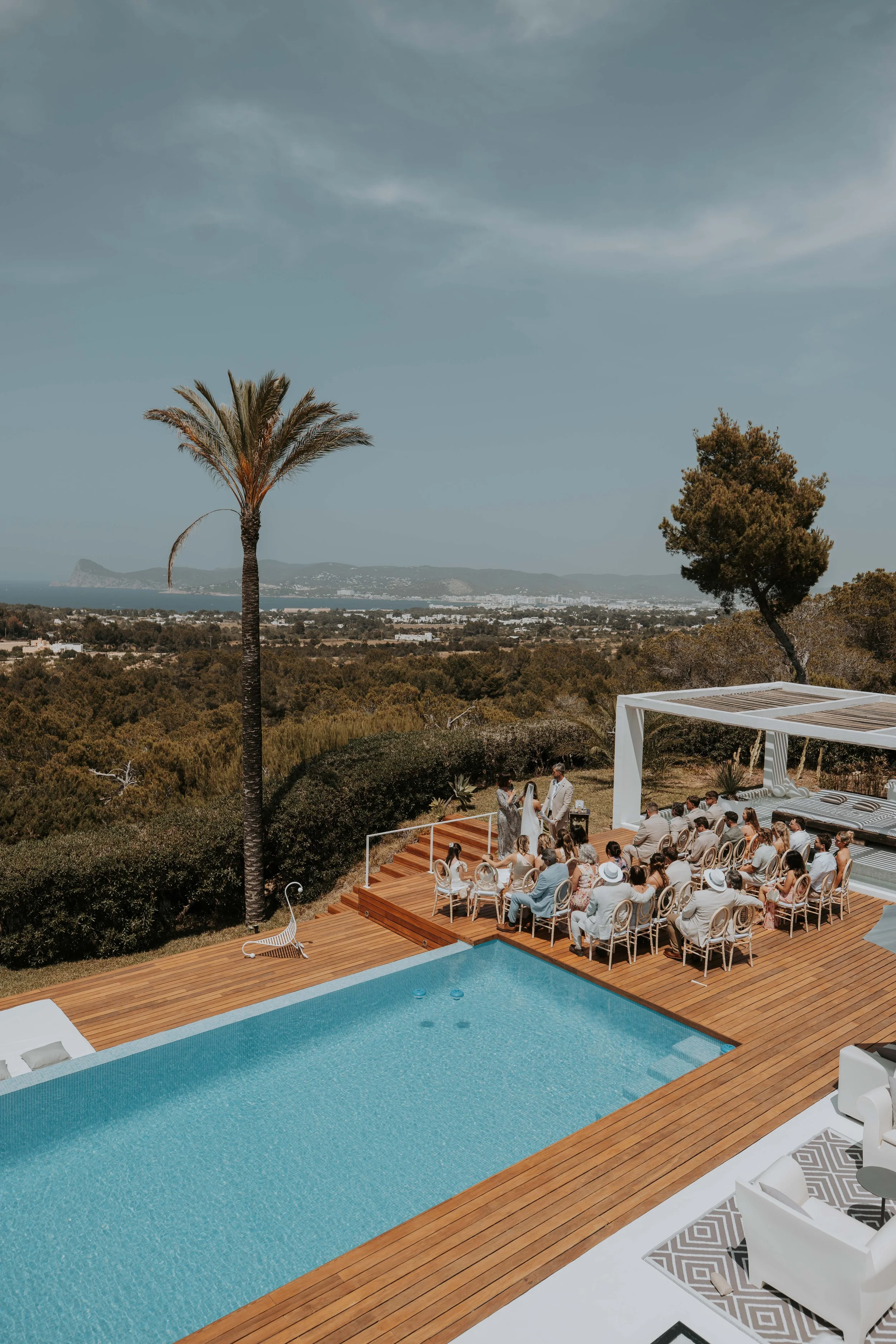 An outdoor wedding ceremony taking place by a swimming pool, with guests seated on chairs on a wooden deck, officiant and bride and groom standing at the altar, in a scenic hillside location with trees and mountains in the background.