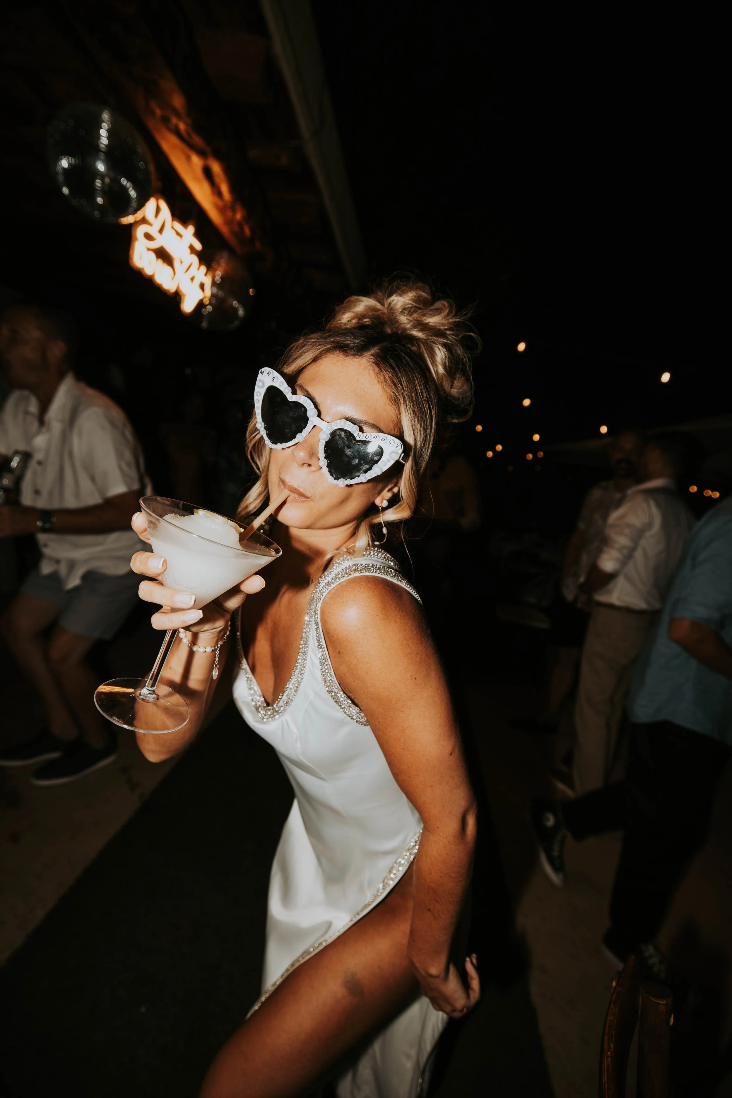 Woman with heart-shaped sunglasses drinking a cocktail at a party or celebration, wearing a white dress with glittery details, in a dimly lit environment.