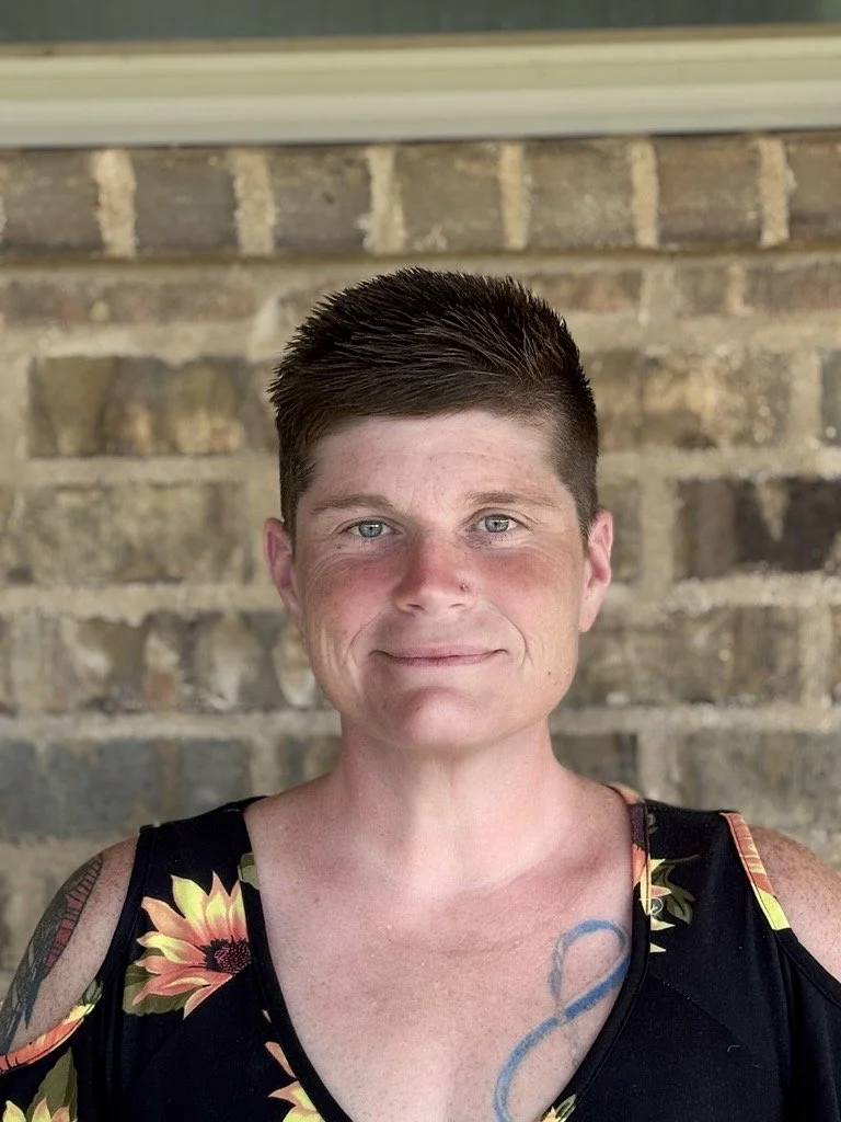A woman with short, dark hair and light skin, smiling slightly. She has a tattoo on her chest and is wearing a black tank top with colorful floral patterns.