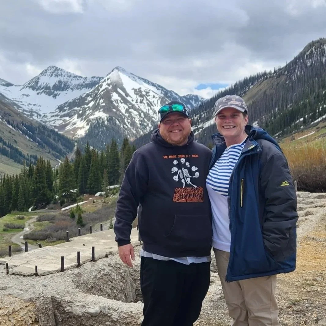Two smiling people stand outdoors in front of snow-capped mountains, green pine trees, and cloudy sky, dressed in outdoor hiking gear.