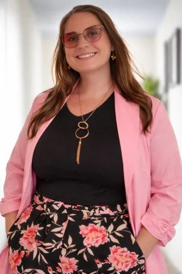 Woman wearing pink blazer, black top, floral skirt, and pink sunglasses, smiling indoors with a blurred background.