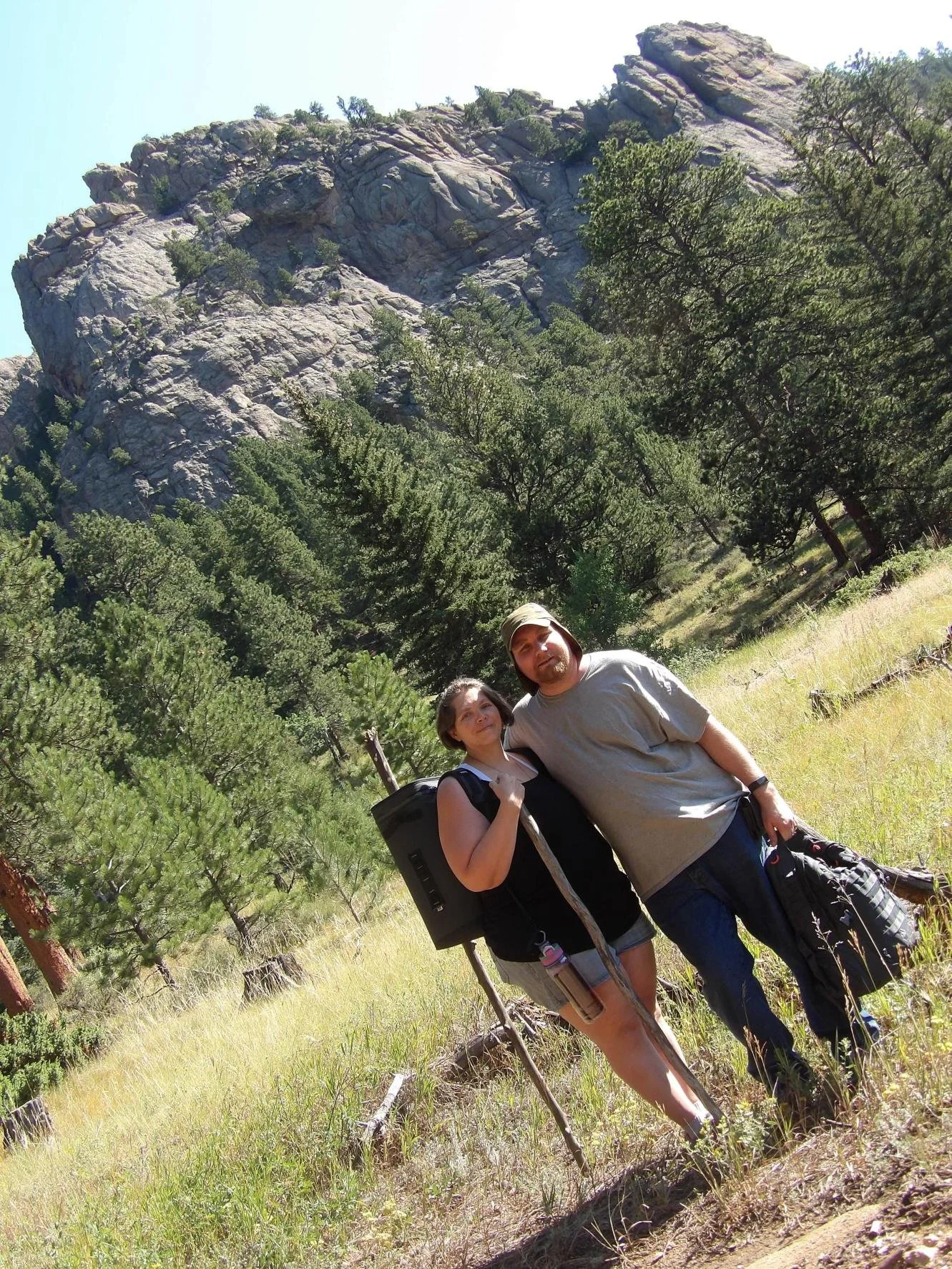 A man and woman hiking in a forested mountainous area with large rocky cliffs in the background, carrying backpacks and hiking sticks.