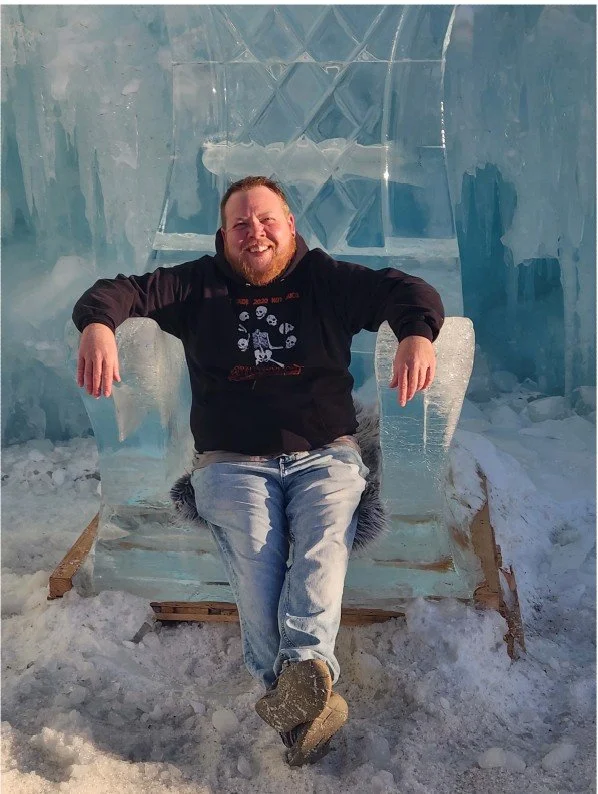 A man sitting on a transparent ice chair inside an ice sculpture room, smiling with his arms resting on the armrests, surrounded by snow.
