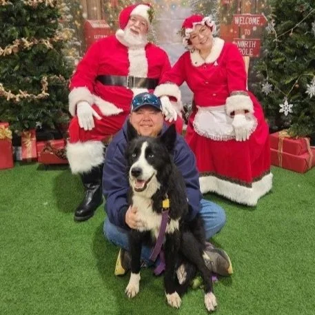 Person kneeling with a black and white dog in front of Santa and Mrs. Claus figures with Christmas decorations.