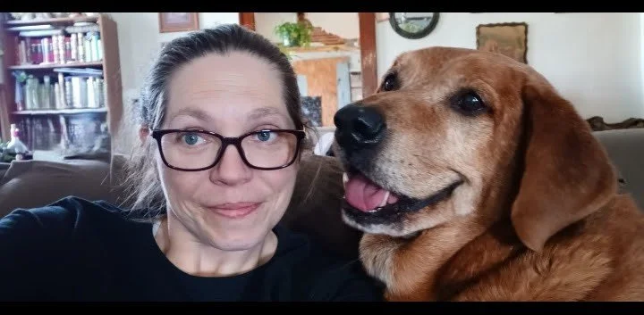 A woman with glasses taking a selfie with her brown dog on a sofa in a cozy living room.