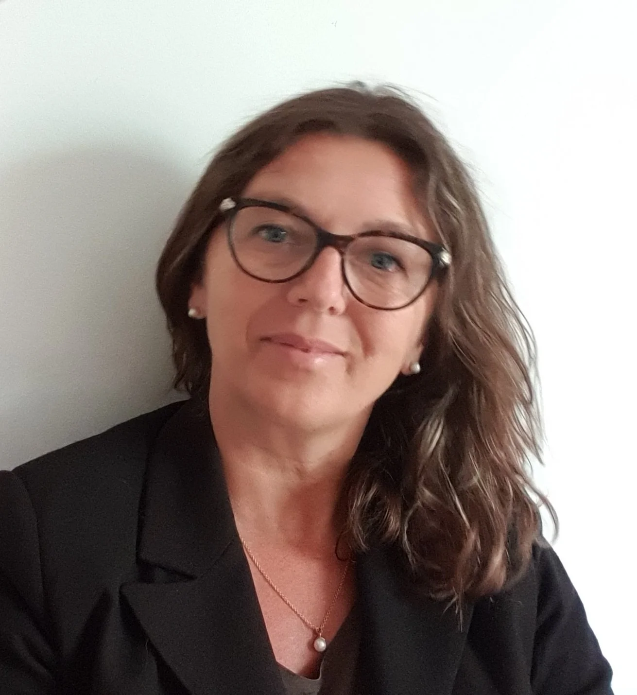 Una donna con capelli mossi e occhiali, indossa un completo nero, poggiata su una parete bianca.