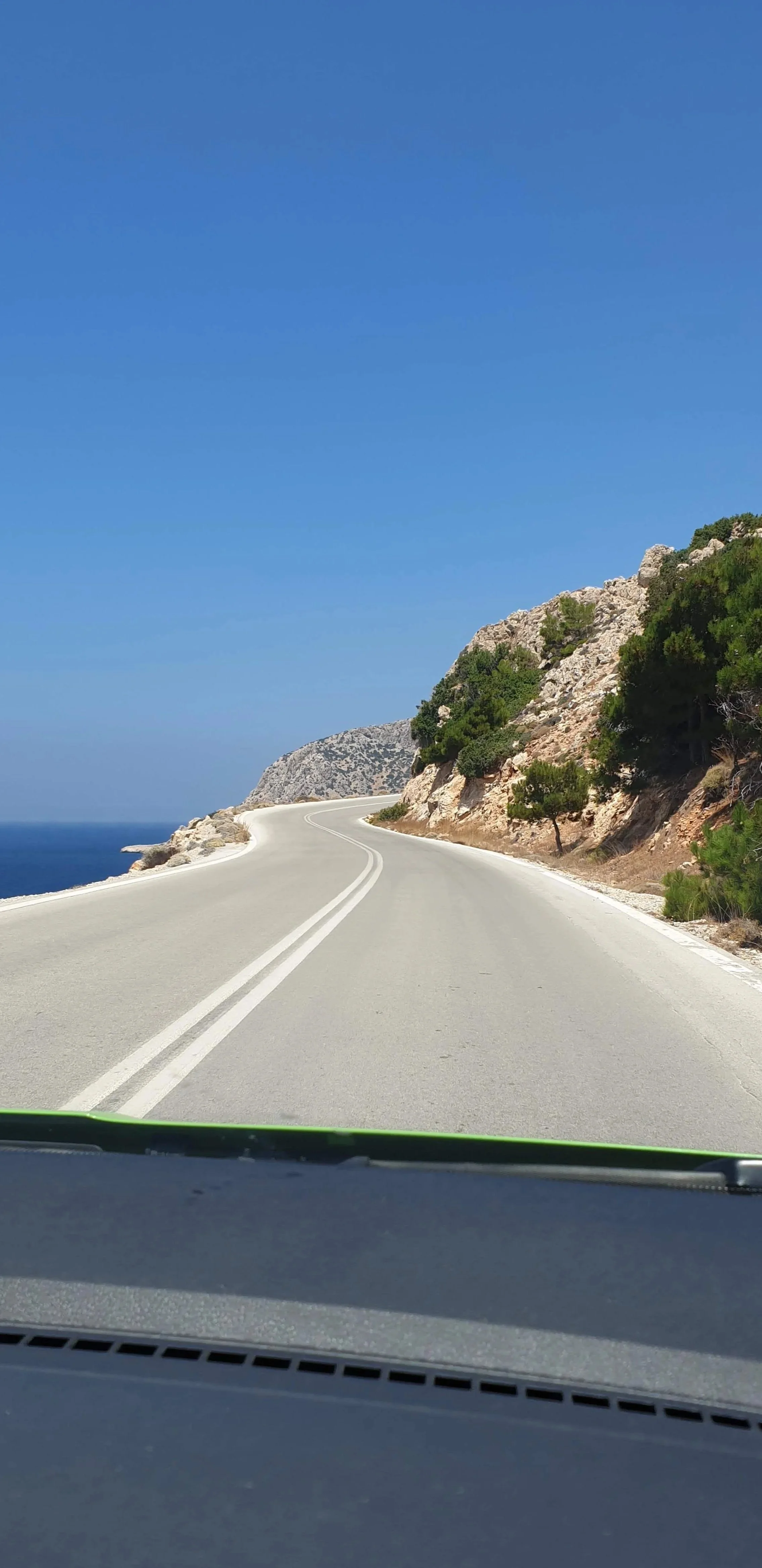 Strada pittoresca costiera con onde sui lati e rocce verdi sotto un cielo azzurro in un paesaggio mediterraneo.