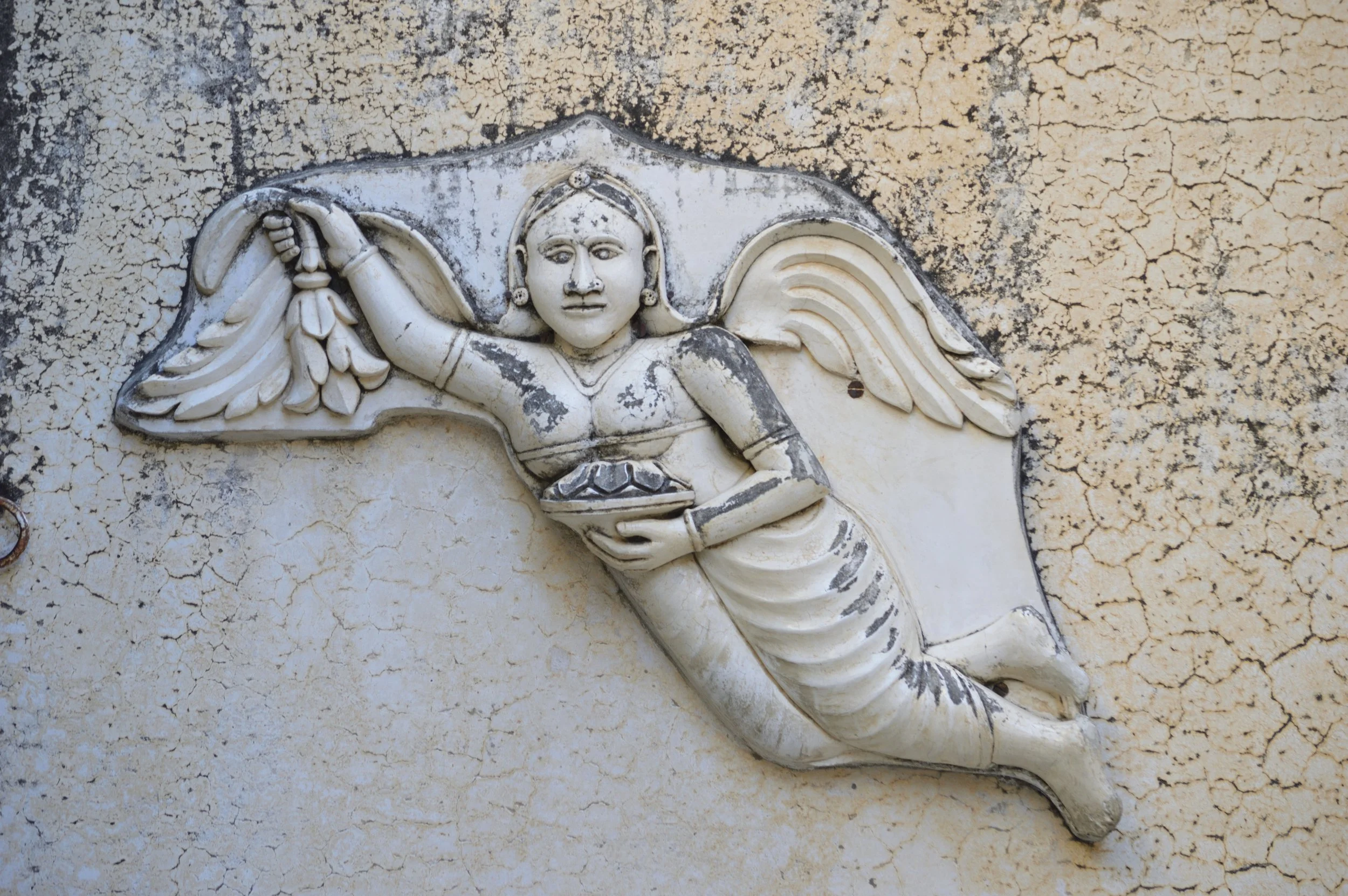 Rilievo di una dea con ali che sorride, che tiene un piatto con foglie, su sfondo di muro crepato.