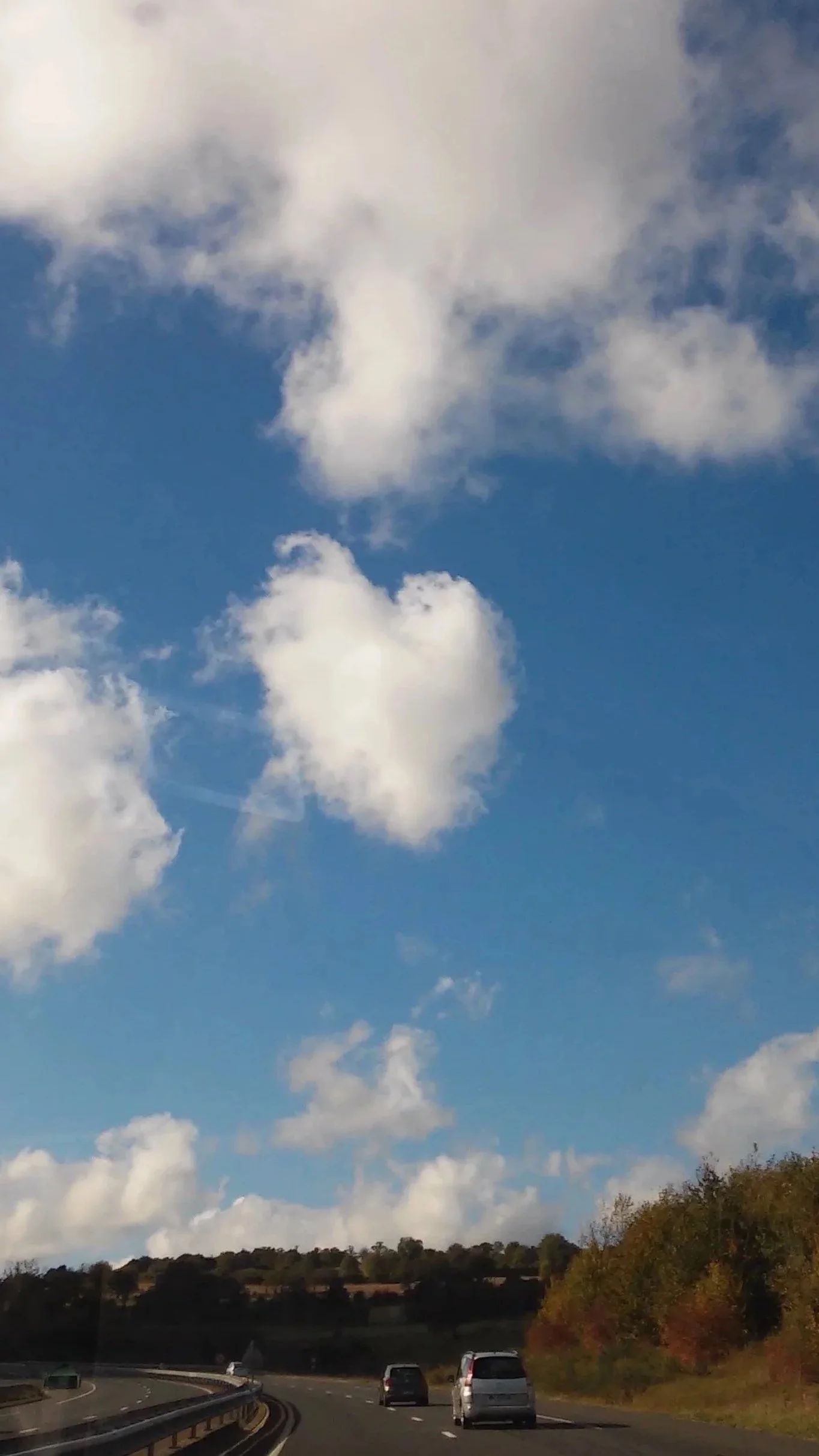 Cielo blu con nuvole bianche a forma di cuore, vista da un'auto in autostrada durante il giorno, con colline e alberi sullo sfondo.