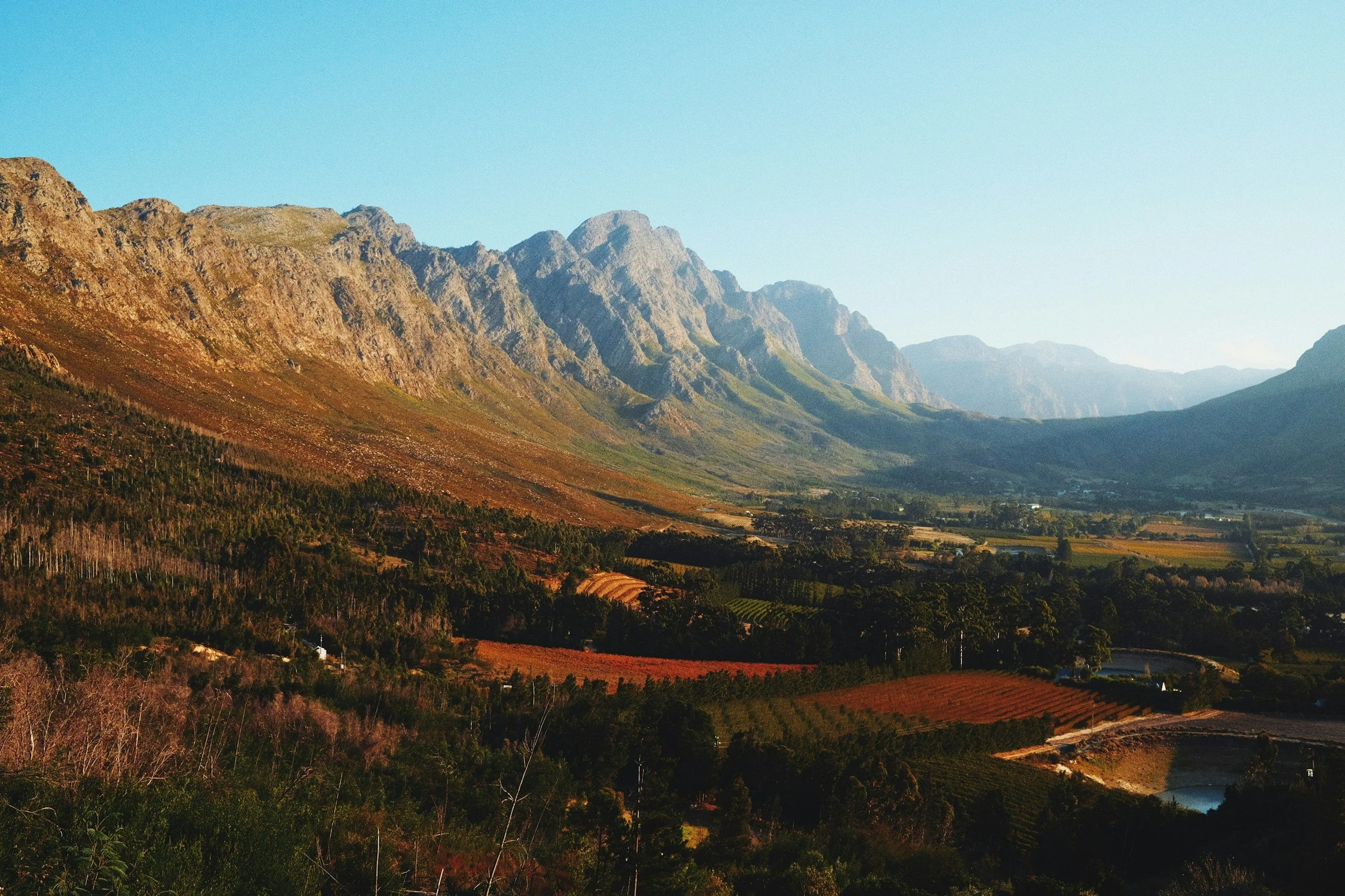 Scenic view of a mountain range with lush valley below, showing green and brown fields, trees, and small water bodies under a clear sky.