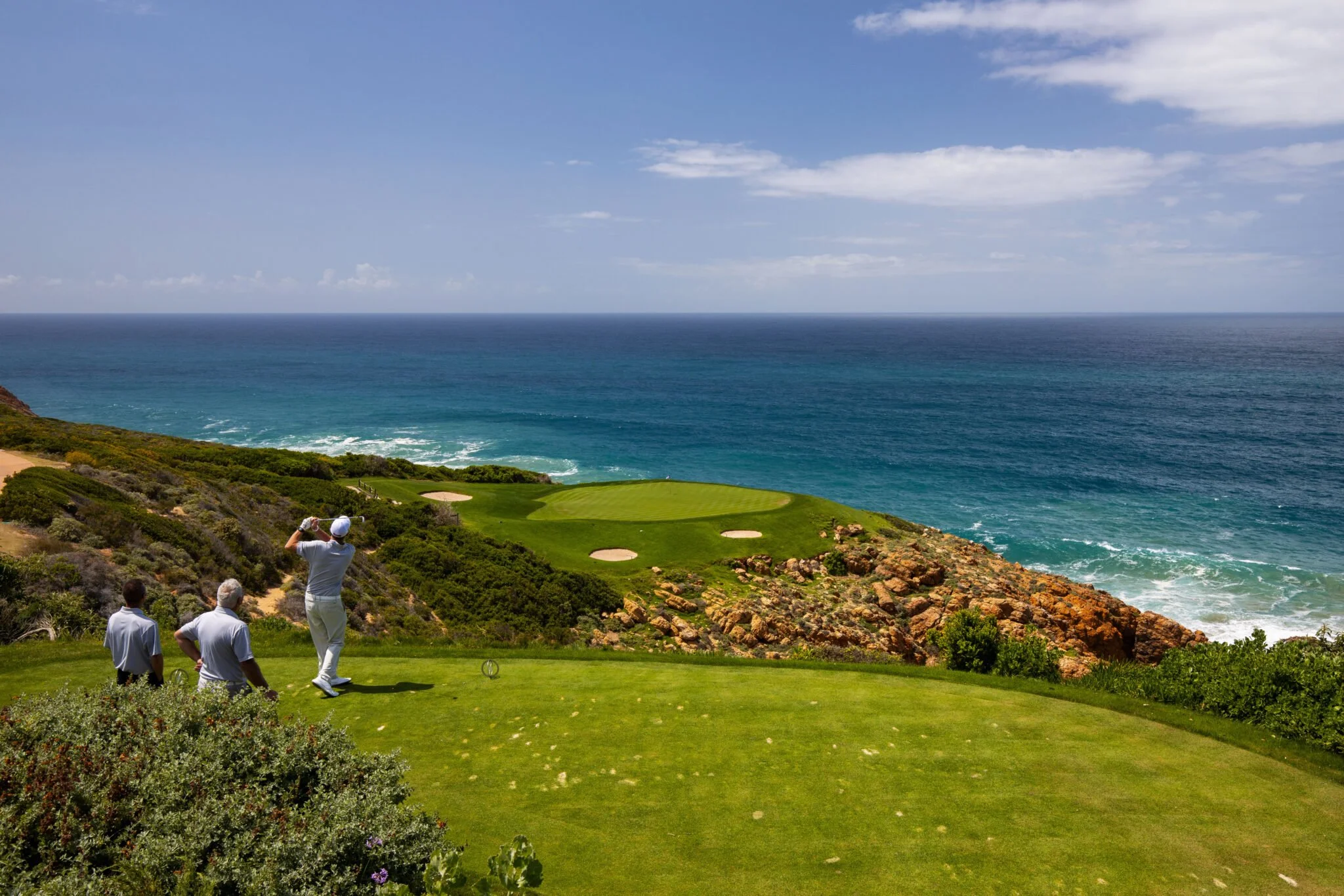 Golfers playing on a course by the ocean, with a green, sand traps, rocky coastline, and blue sky.